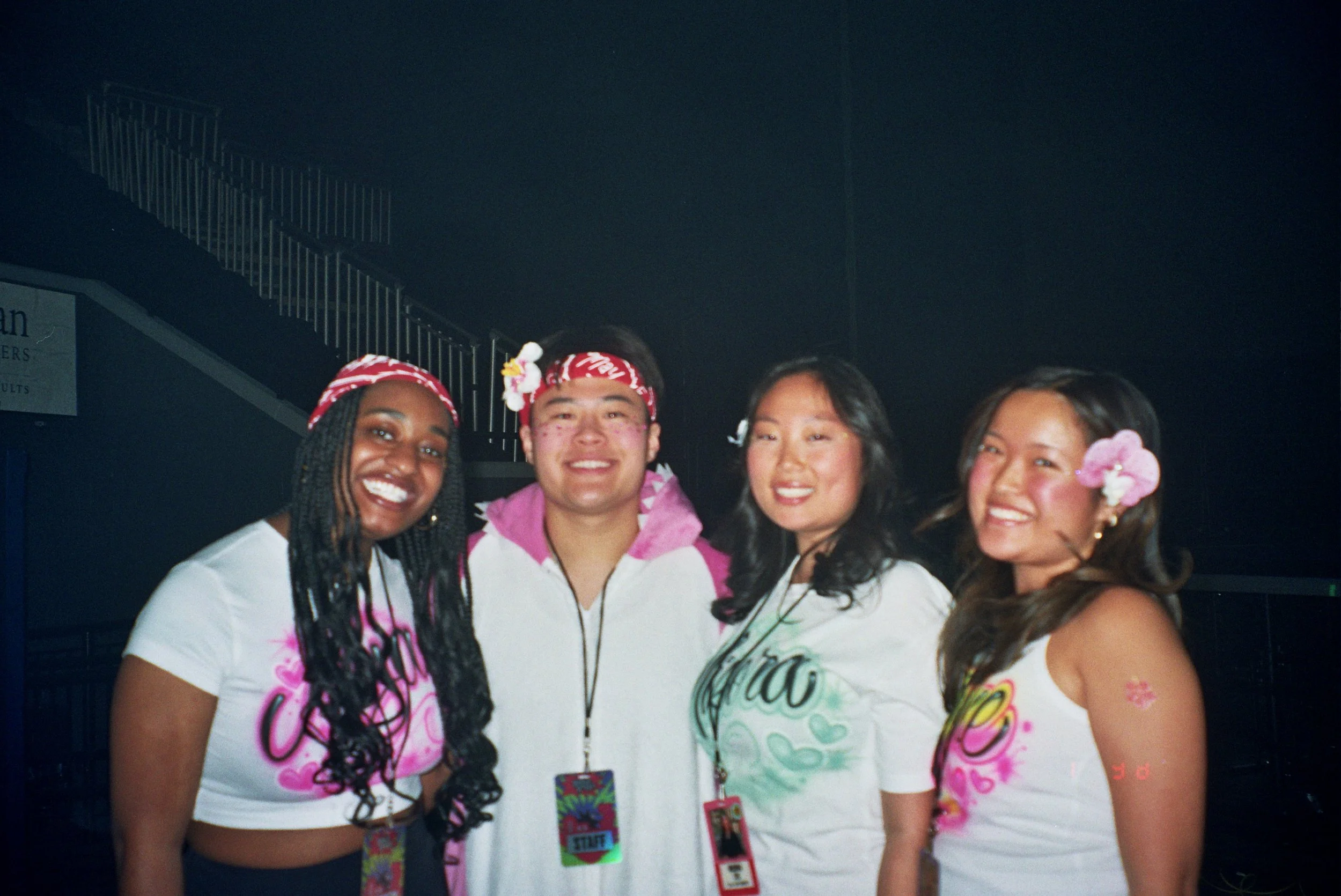 Group of diverse smiling young women wearing white T-shirts with colorful designs, some with pink or floral accessories, standing together indoors.