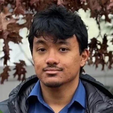 A young man with dark hair and a slight mustache, wearing a blue shirt and a dark jacket, standing outdoors with trees in the background.