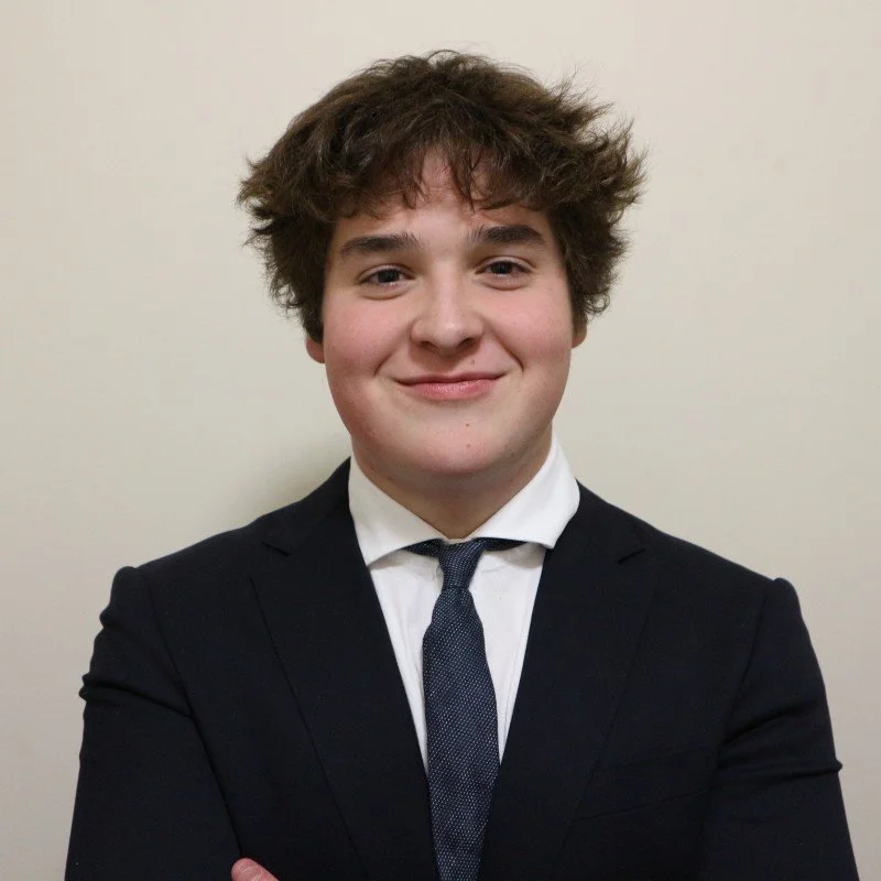 A young man with curly brown hair wearing a black suit, white shirt, and dark tie, smiling and standing against a plain beige background.