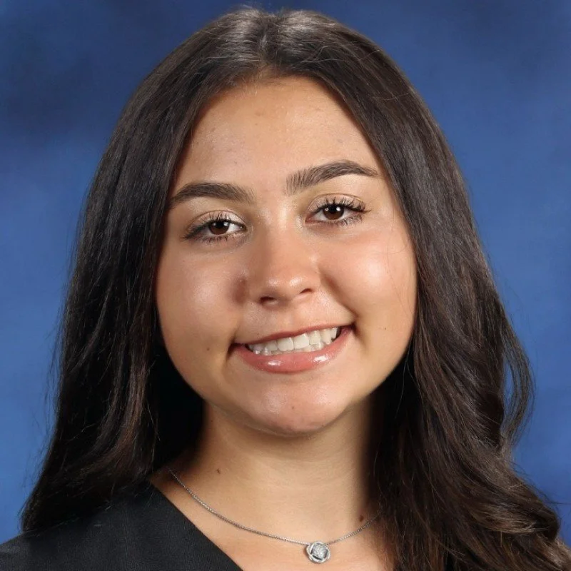 A young woman with long dark hair, smiling, wearing a necklace, and a black top against a blue background.