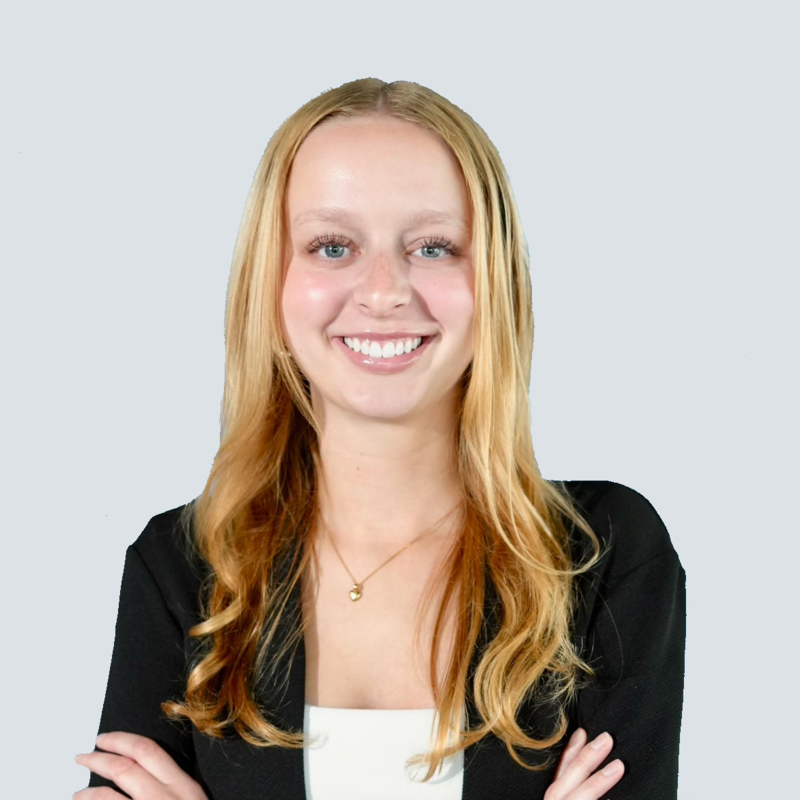 A young woman with long red hair, blue eyes, and fair skin smiling with arms crossed, wearing a black blazer and a white top against a light gray background.