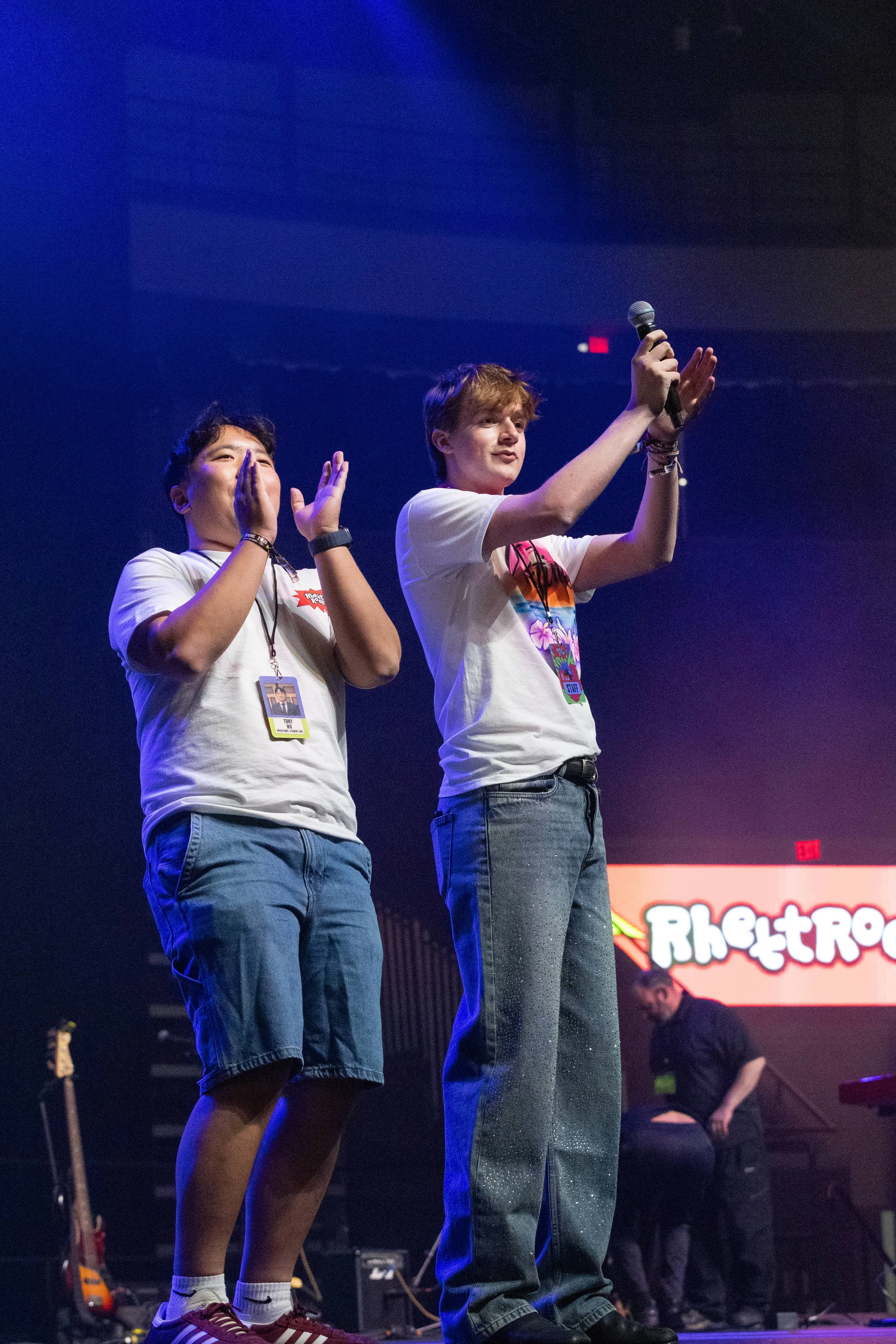 Two young men on stage, one holding a microphone, wearing casual clothing and jeans, with a bright screen behind them and musical instruments on stage.