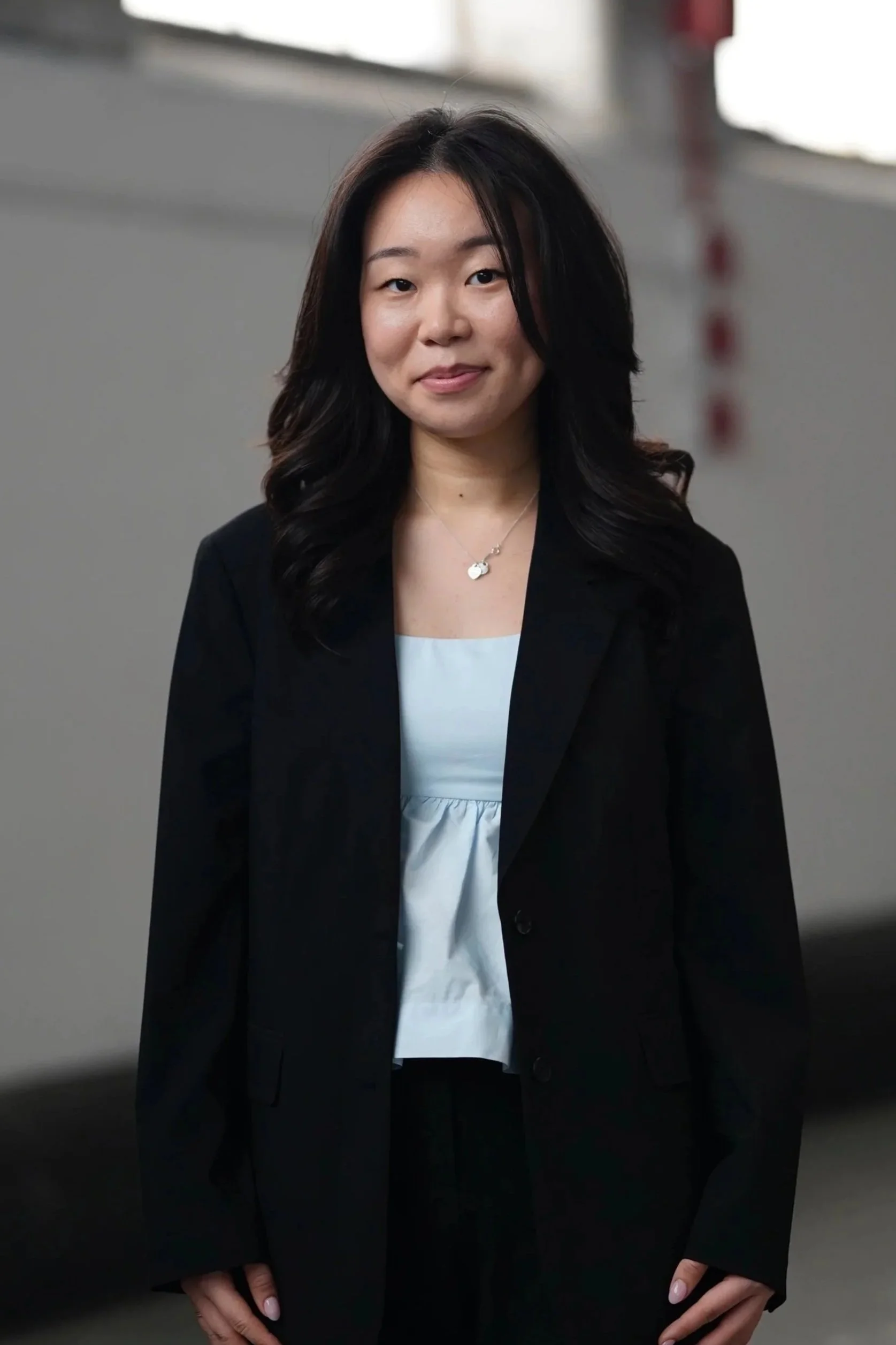 A woman with long dark hair wearing a black blazer, white top, and a necklace, standing in a parking garage.