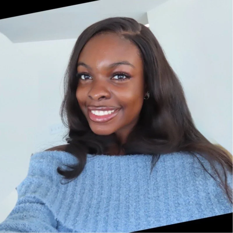 A young woman with long dark hair, smiling while taking a selfie indoors, wearing a light blue sweater.