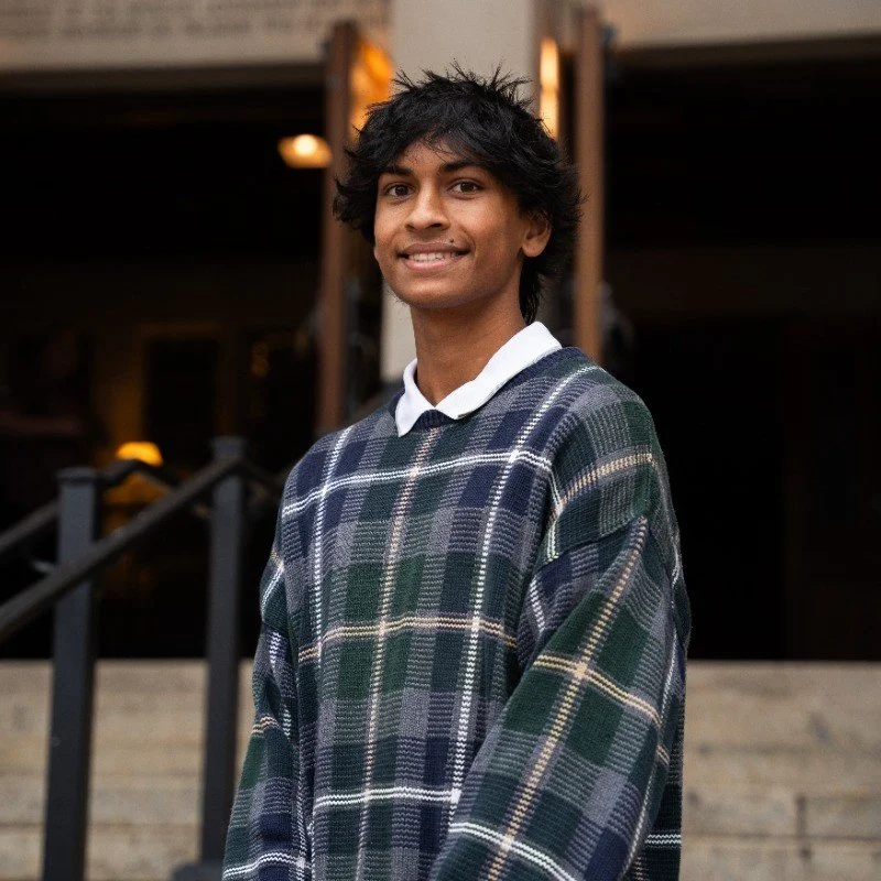 Young man with dark hair wearing a checkered sweater over a white collared shirt stands outdoors in front of a building.