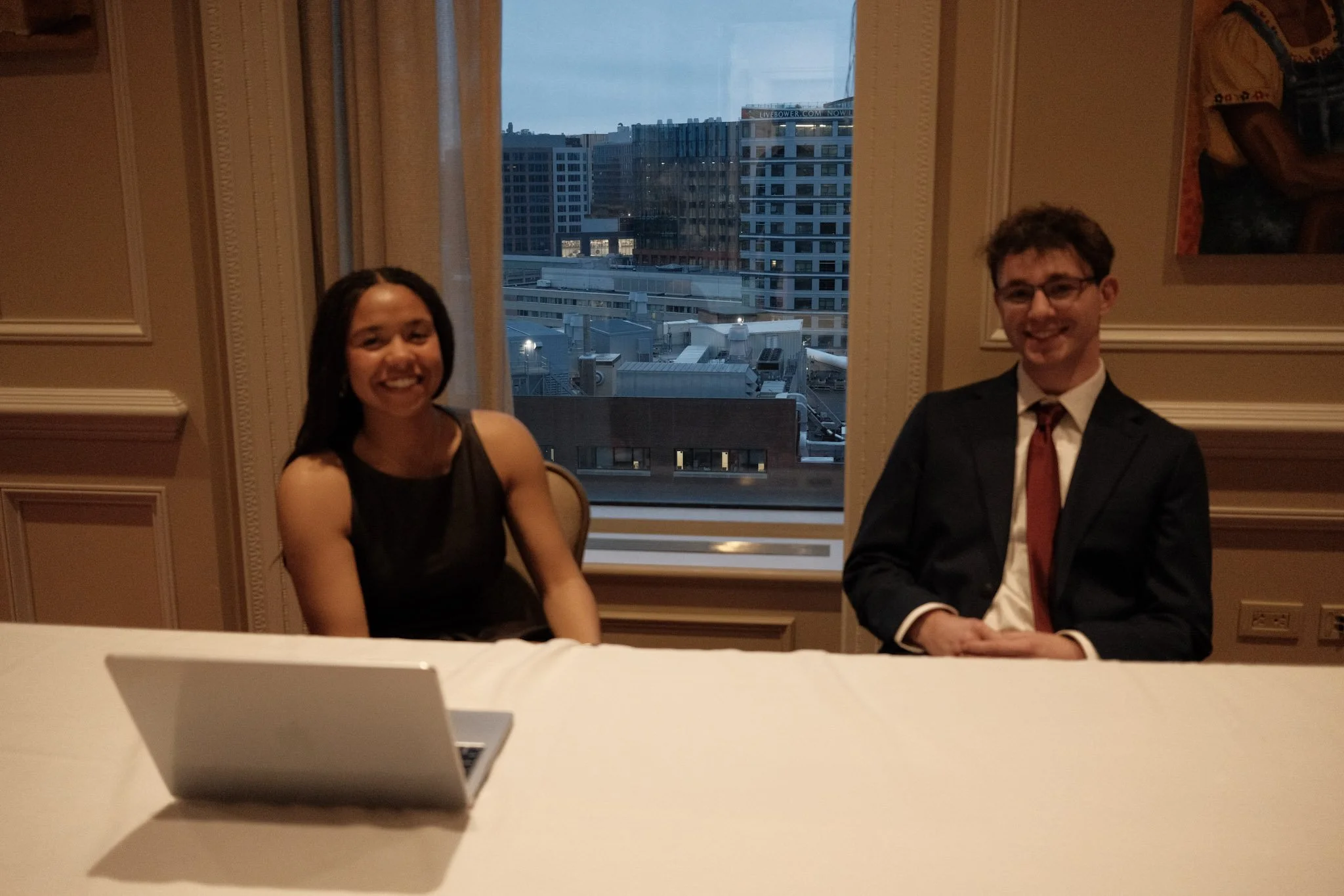 A woman and a man sitting at a table in front of a window with a view of city buildings during evening. The woman is smiling, wearing a black dress. The man is smiling, wearing a suit and tie, and has glasses.