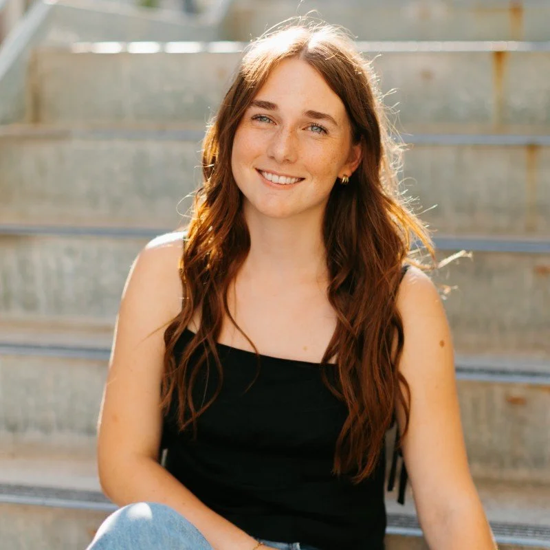 A young woman with long wavy auburn hair, blue eyes, and freckles, wearing a black tank top, smiling outdoors on metal stairs with sunlight shining on her.