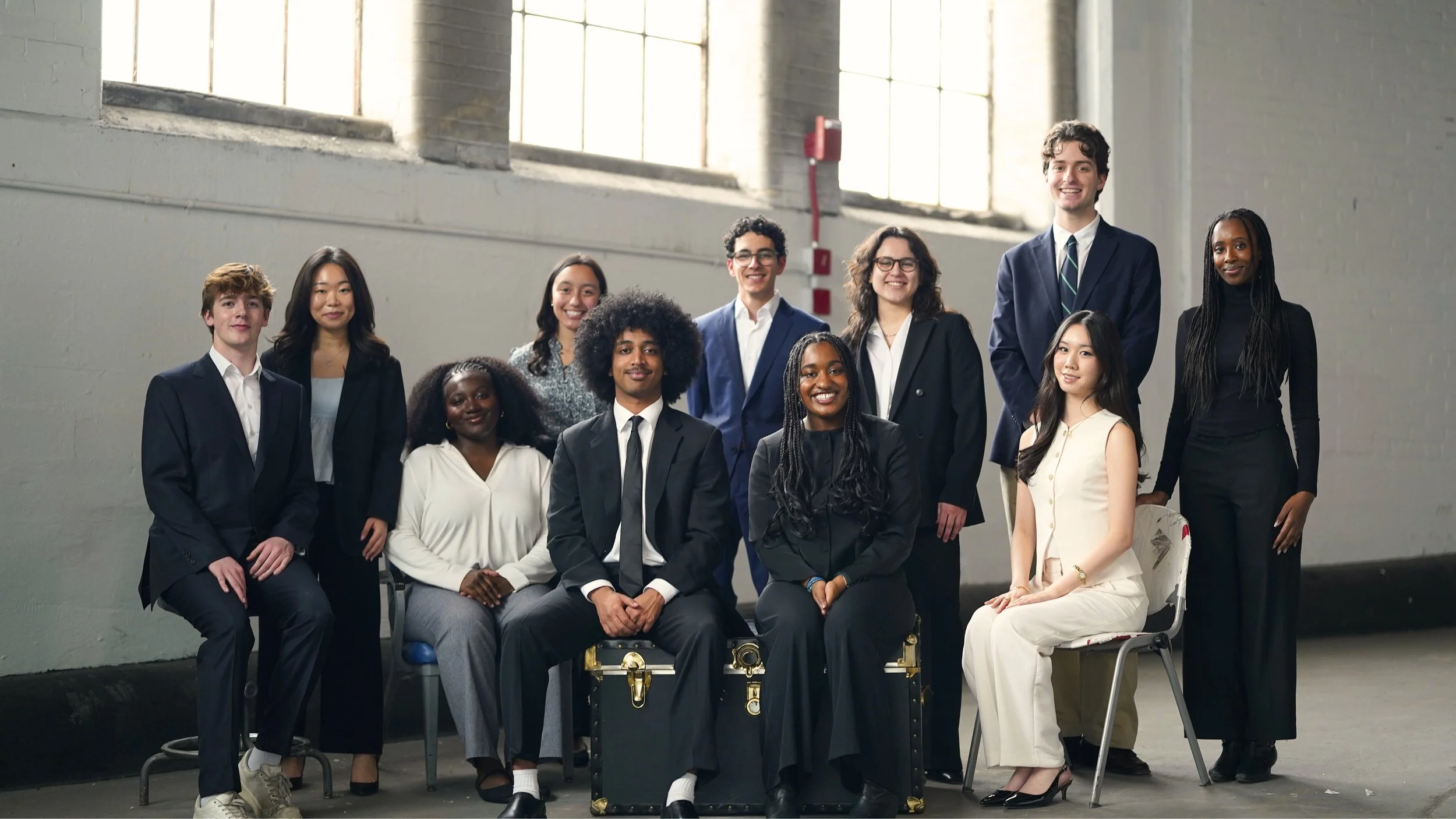 Group of diverse young professionals posing together in an industrial-style space with large windows, dressed in business and smart casual attire.