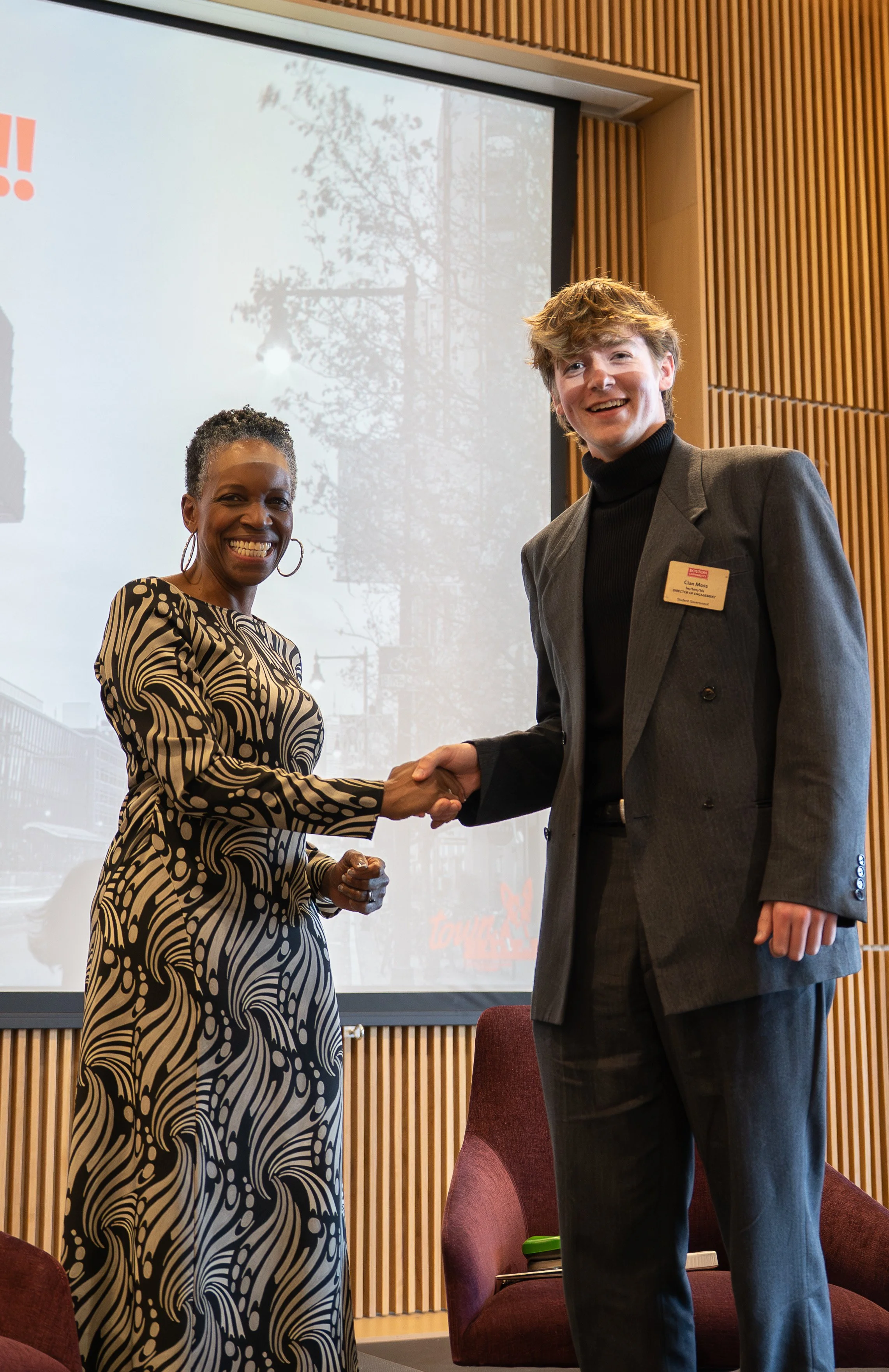 Two people shaking hands at an indoor event, with a woman on the left and a man on the right. The woman is wearing a black and white patterned dress, and the man is in a gray blazer with a name badge. They are smiling and standing in front of a presentation screen and wood-paneled wall.