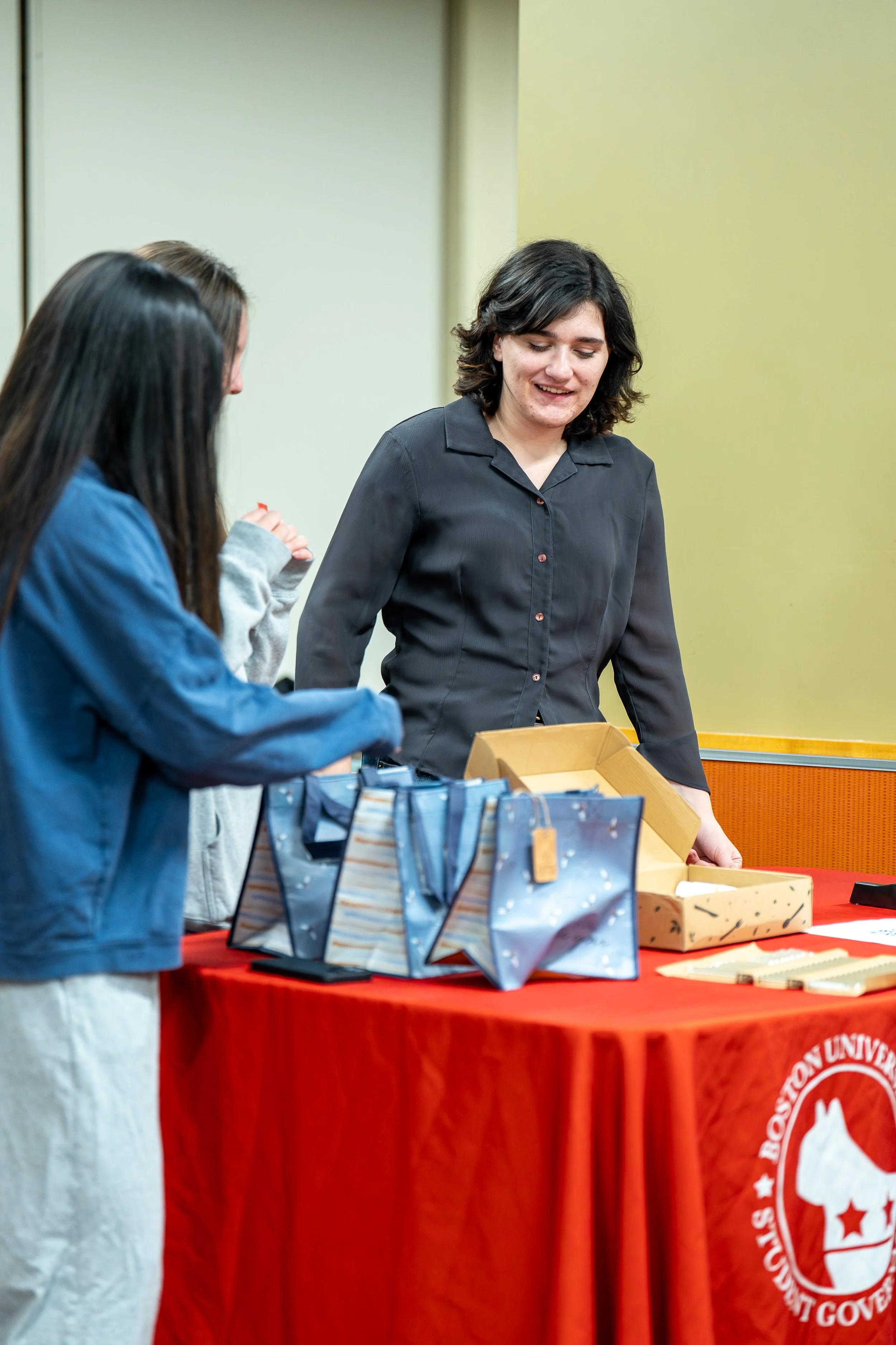 A woman with dark, wavy hair and a black shirt is standing behind a table with a red tablecloth. There are paper bags with blue and white patterns on the table. The woman is smiling and looking down at the table. The table has a logo that reads "Borough of the United States" with a dog silhouette in the center.
