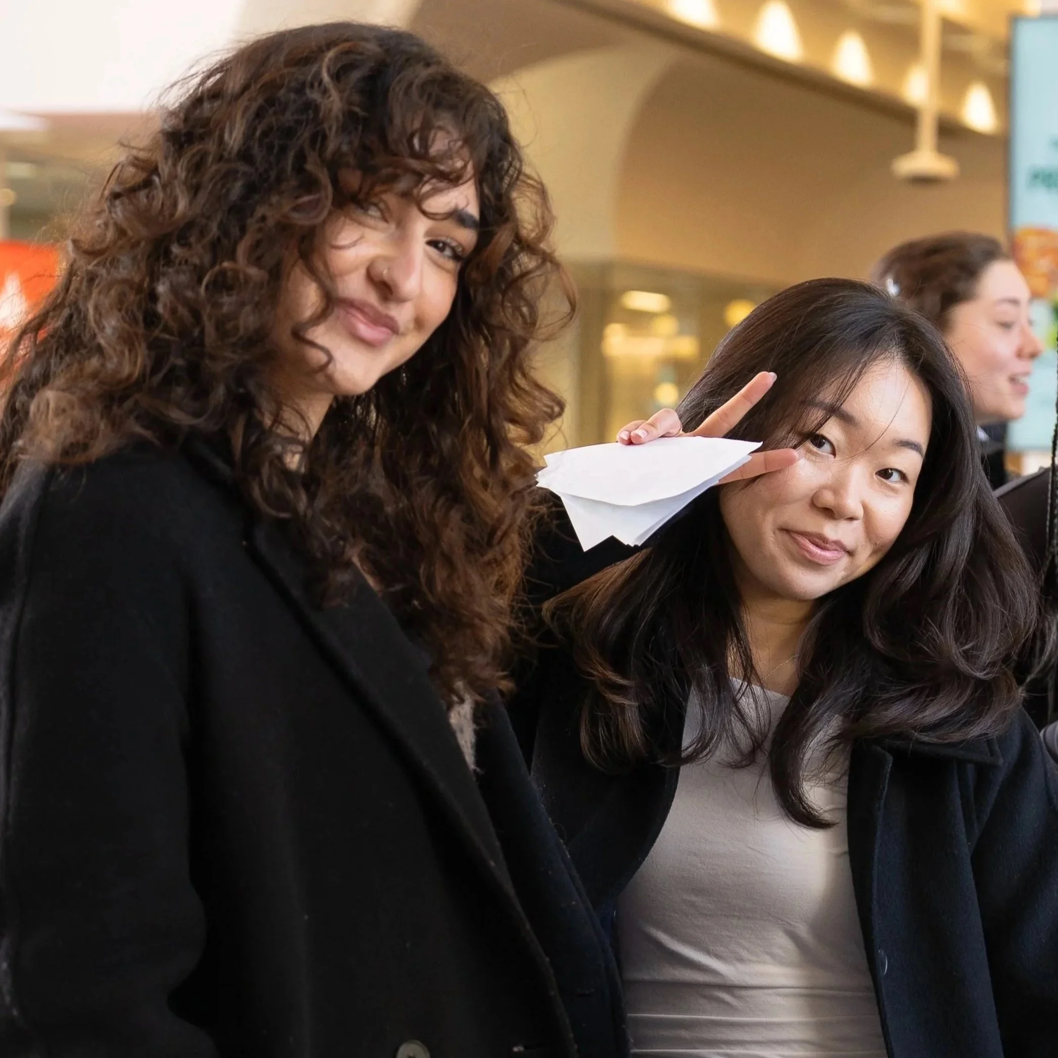 Two young women with long dark hair, one with curly hair, are close to each other indoors. One woman is making a peace sign and appears to be smiling, while the other is looking slightly downward. The background shows a blurred supermarket or shopping mall.