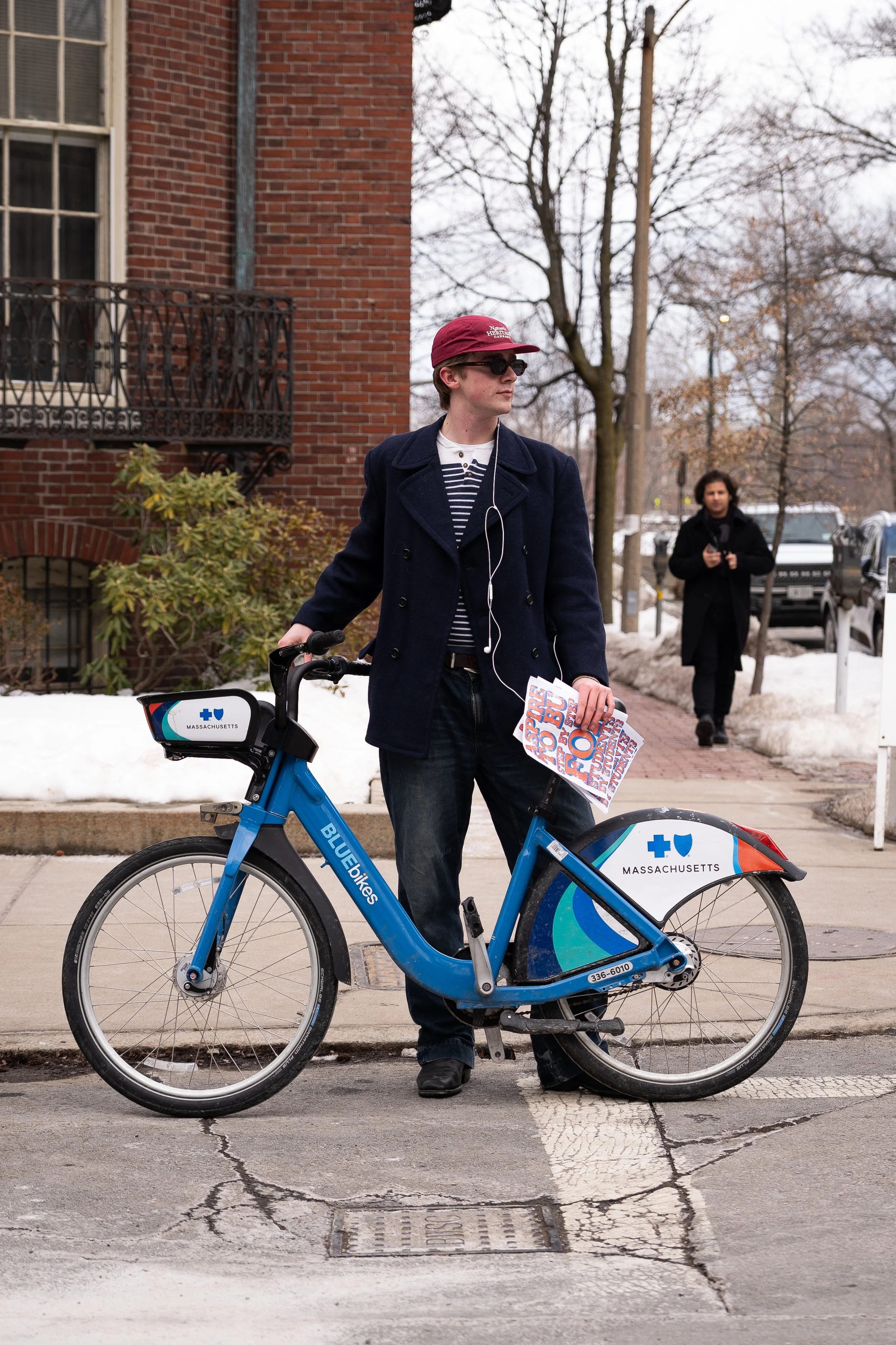 A person wearing sunglasses, a red baseball cap, a navy coat, and dark pants stands next to a Bluebikes bicycle on a city street. They are holding a newspaper and have earphones in, and are looking to their left. In the background, another person is walking, dressed in dark clothing, with trees and parked cars on a snowy day.