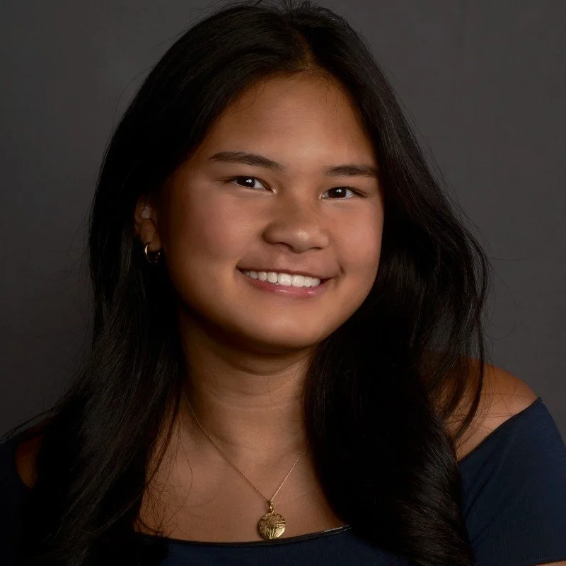 A young woman with long dark hair, smiling, wearing a dark off-the-shoulder top, a gold necklace with a round pendant, and small hoop earrings, against a dark background.