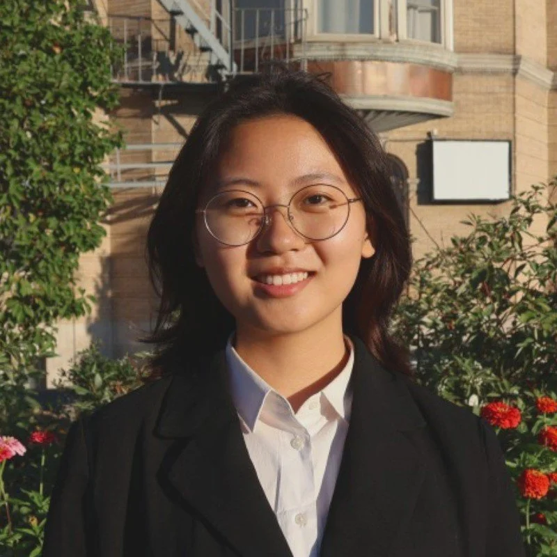 A young woman with glasses, wearing a white shirt and black blazer, smiling outdoors with a building and plants in the background.