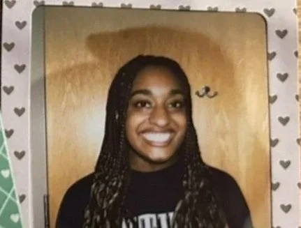 A young woman with braids smiling at the camera, standing in front of a wooden door with a heart-patterned border around the photo.