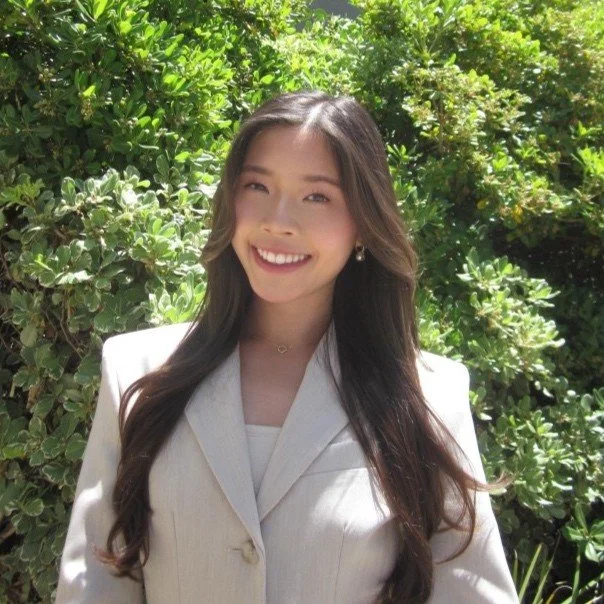 Young woman outdoors with green foliage, smiling and winking, wearing a light-colored blazer and small earrings.