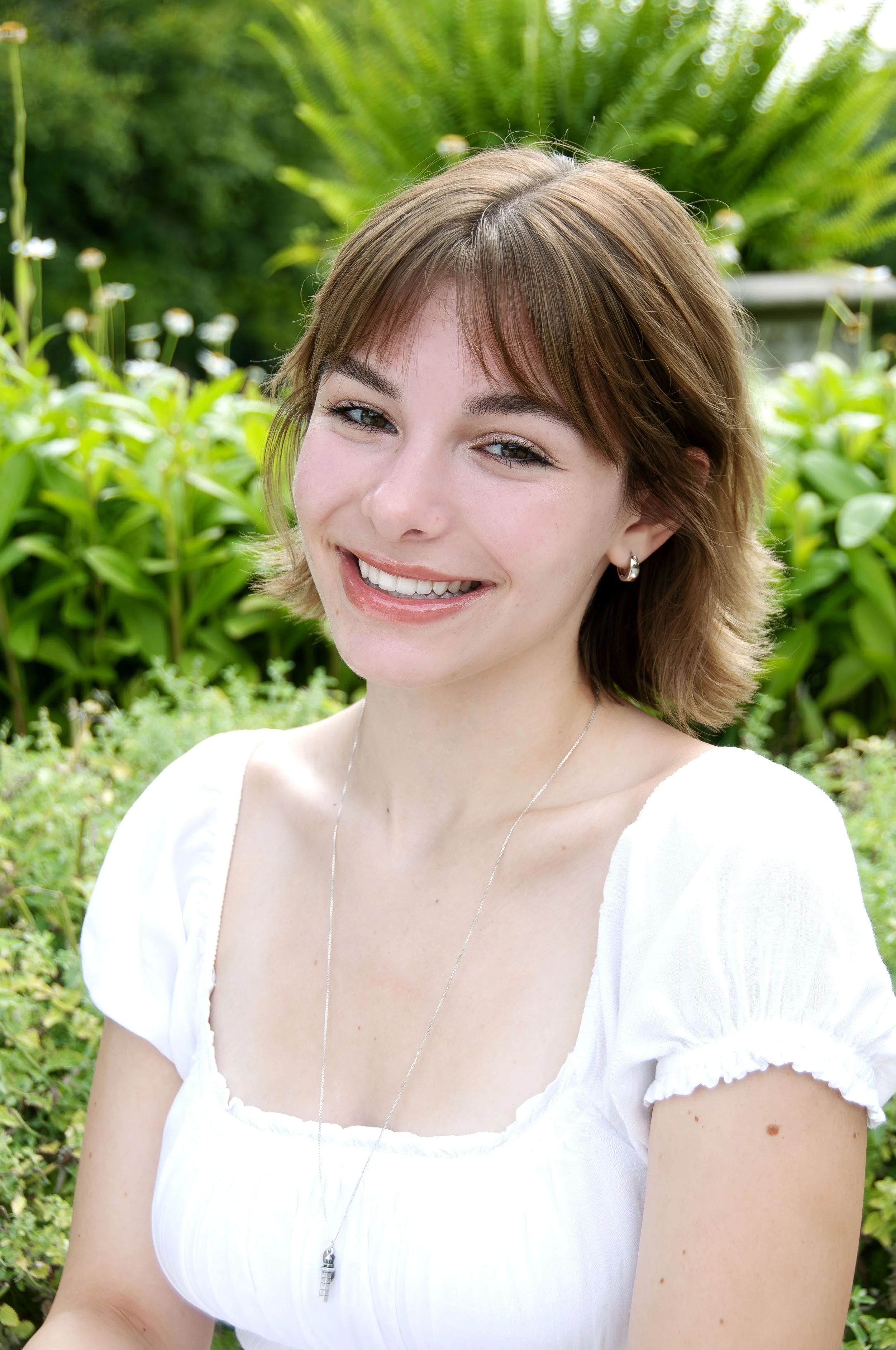 A smiling woman with short brown hair and earrings, wearing a white dress with ruffled sleeves, standing outdoors with greenery in the background.