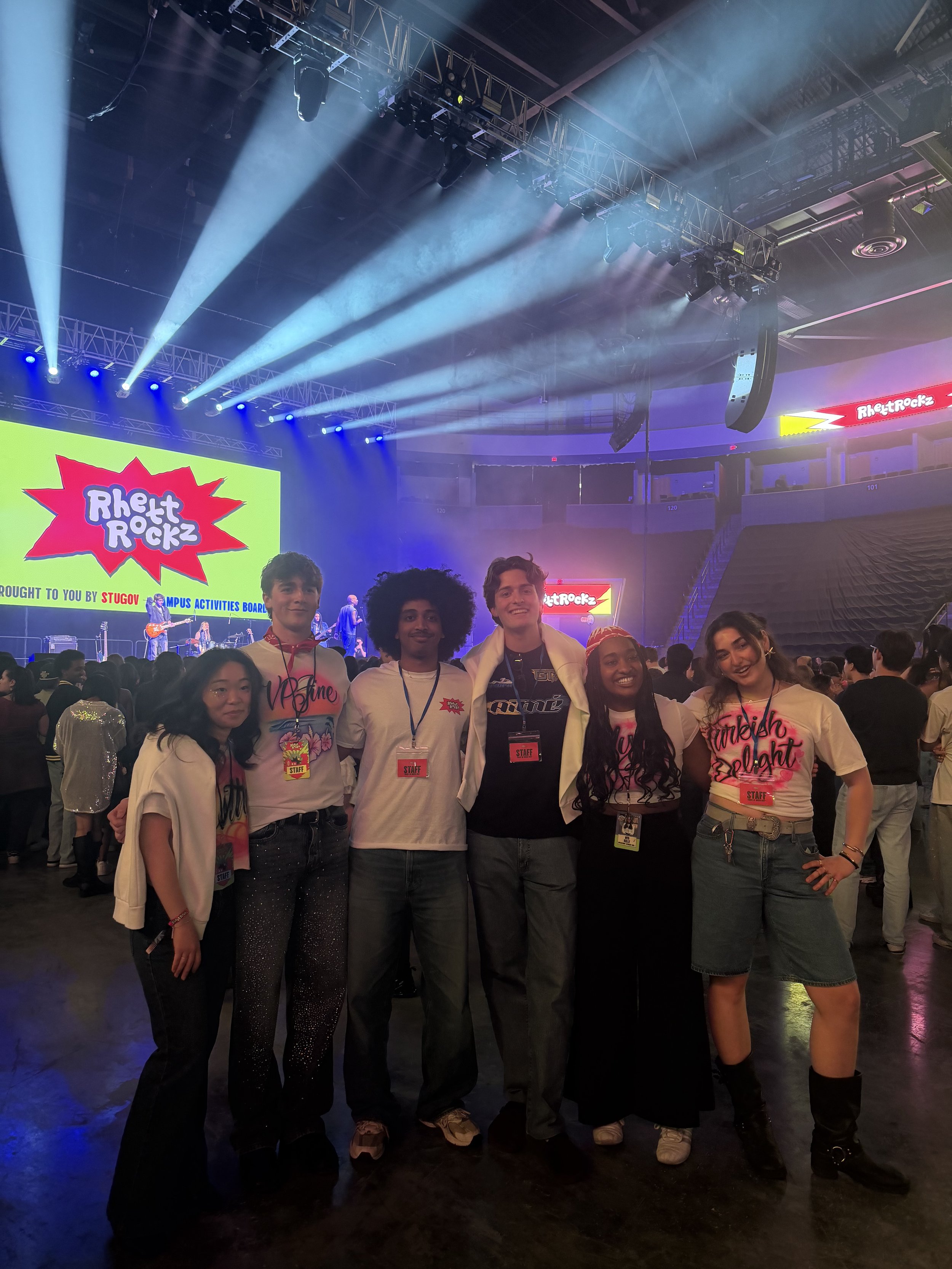 A group of six young people standing together at an indoor concert or event, with a stage, lights, and a large screen displaying 'Rhett Ruckz' in the background.