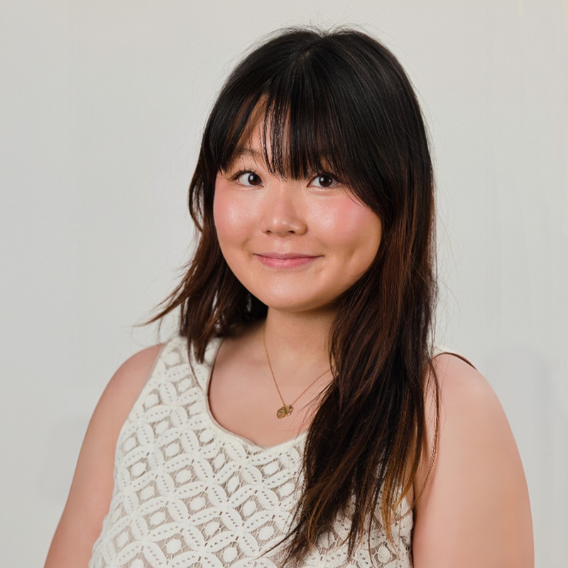 A young woman with shoulder-length dark hair and bangs, smiling gently, wearing a cream-colored crochet sleeveless top and a delicate necklace, standing against a plain white background.