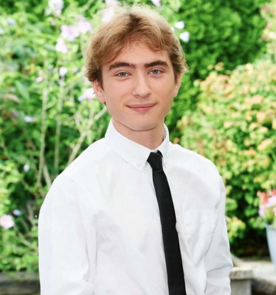 A young man with light brown hair and blue eyes wearing a white dress shirt and black tie, standing outdoors with green foliage in the background.