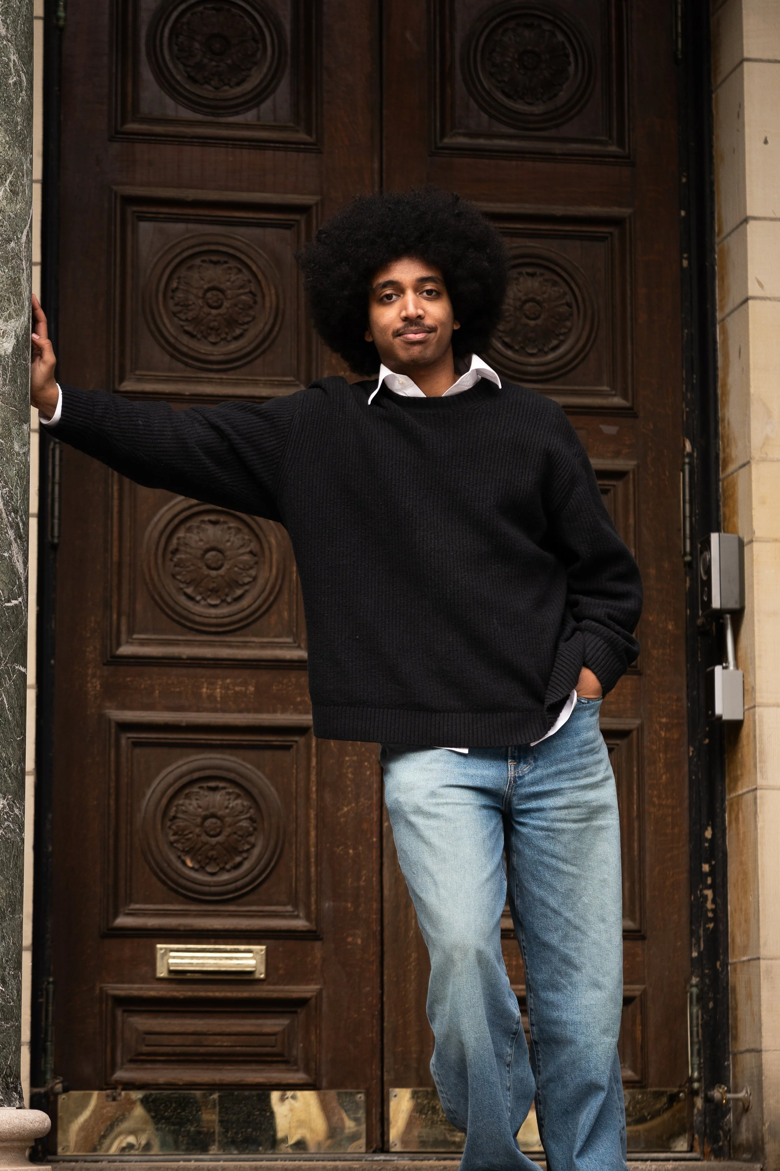 A young man with an afro hairstyle, wearing a black sweater over a white shirt and blue jeans, standing in front of a large wooden door with decorative carvings.