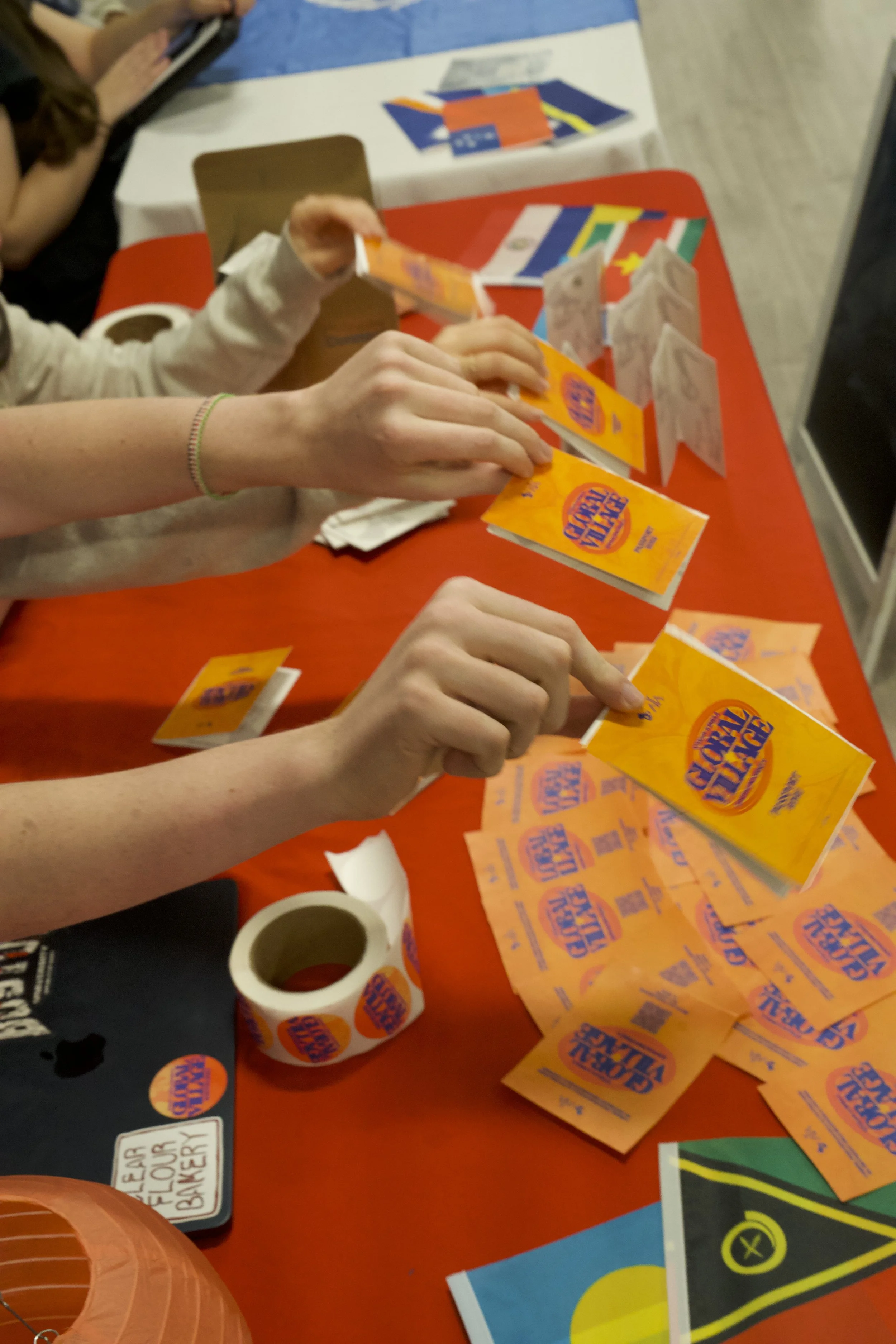 People folding flyers or pamphlets with orange and blue text on a red table, with rolls of stickers nearby, in an indoor setting.