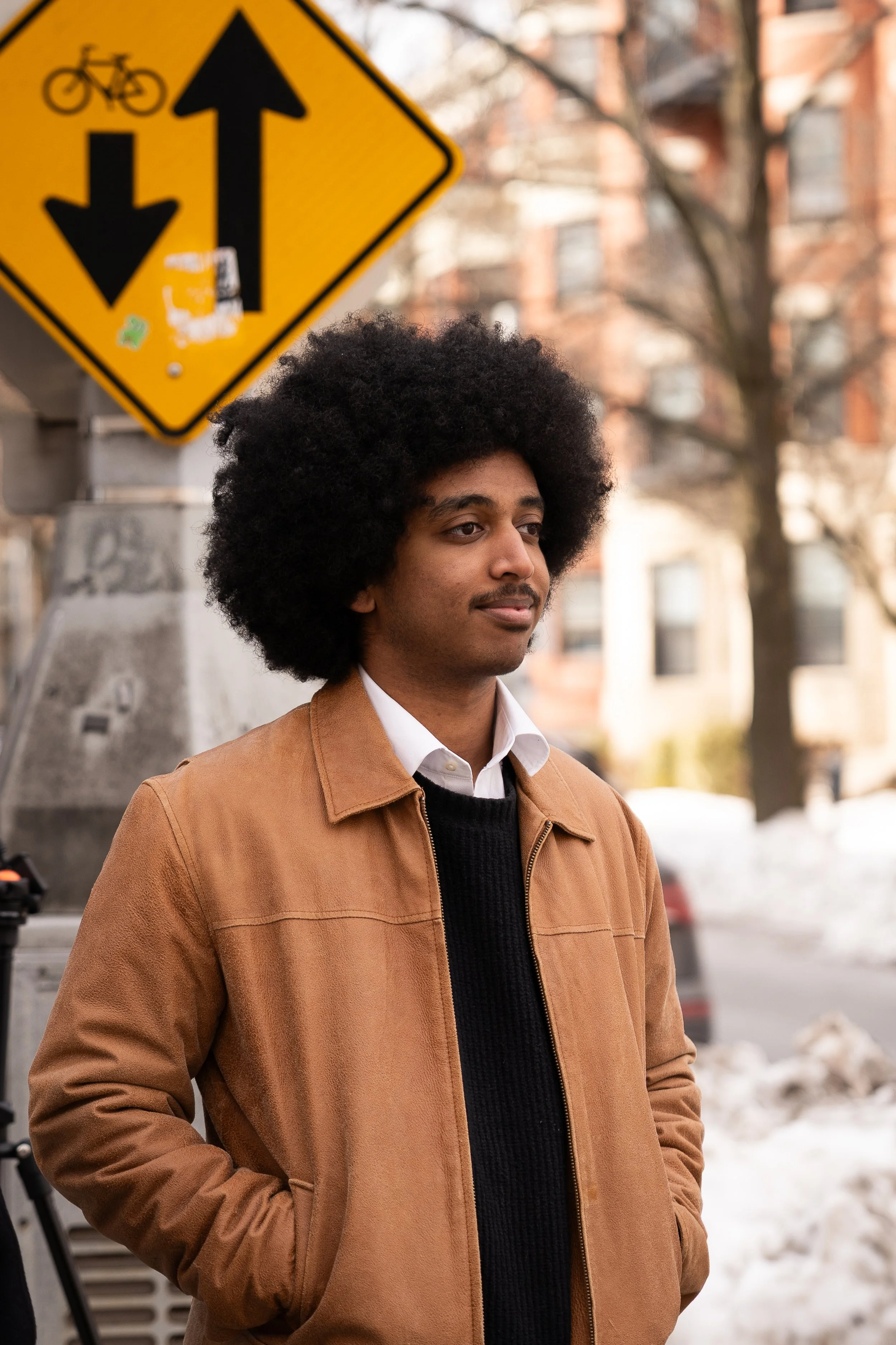 A young man with an afro hairstyle standing outdoors near a building with a yellow street sign in the background that depicts a bicycle and arrows pointing in opposite directions. There are trees and snow on the ground.