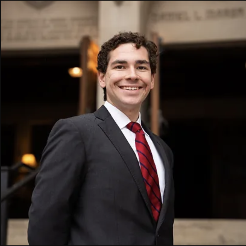 A smiling man wearing a black suit, white shirt, and red striped tie standing outside at night.