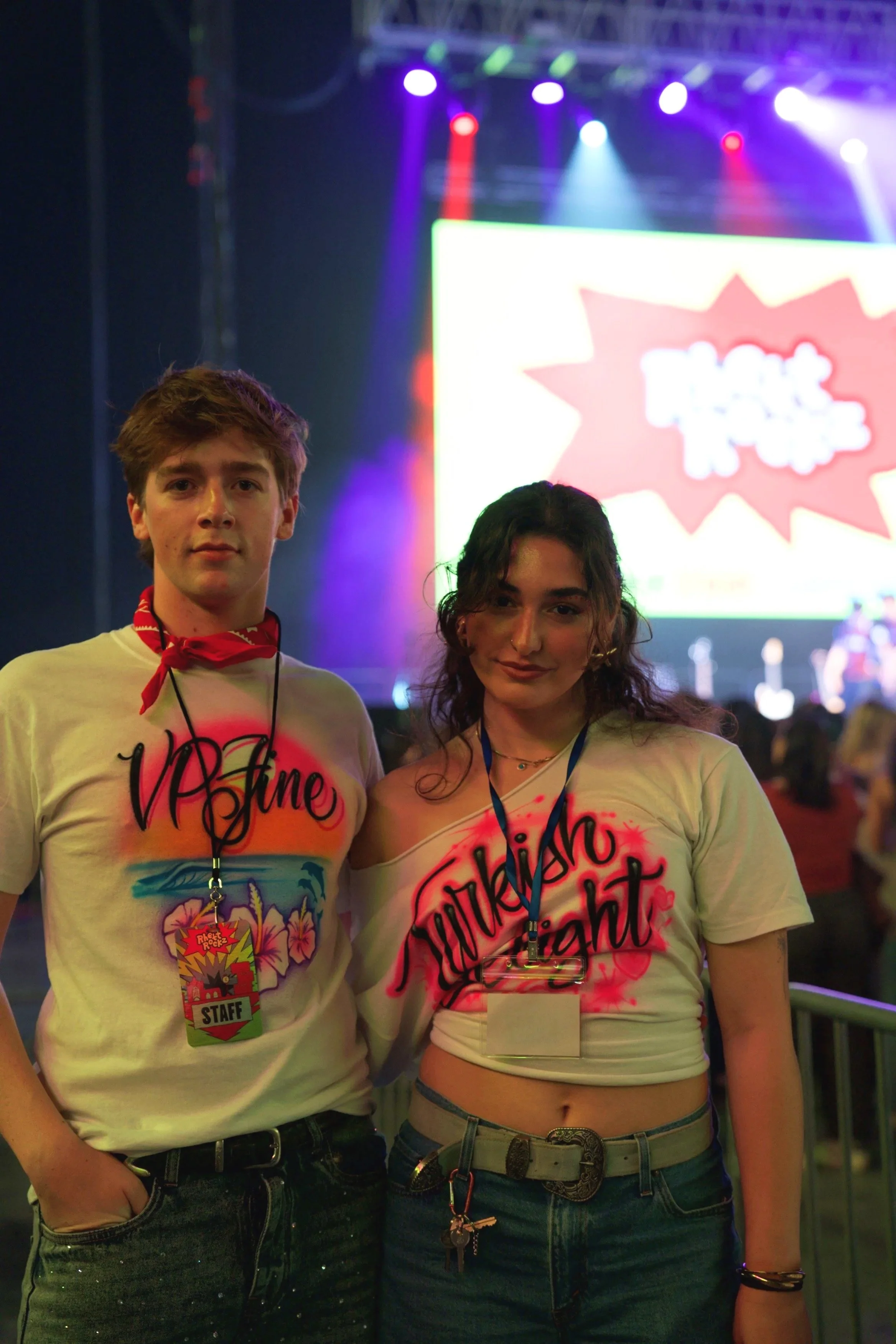 Two young women standing together at a lively event or concert. They are wearing matching white crop tops with colorful, bold text. Behind them, there is a stage with purple and blue lights, and a large screen displaying vibrant graphics.