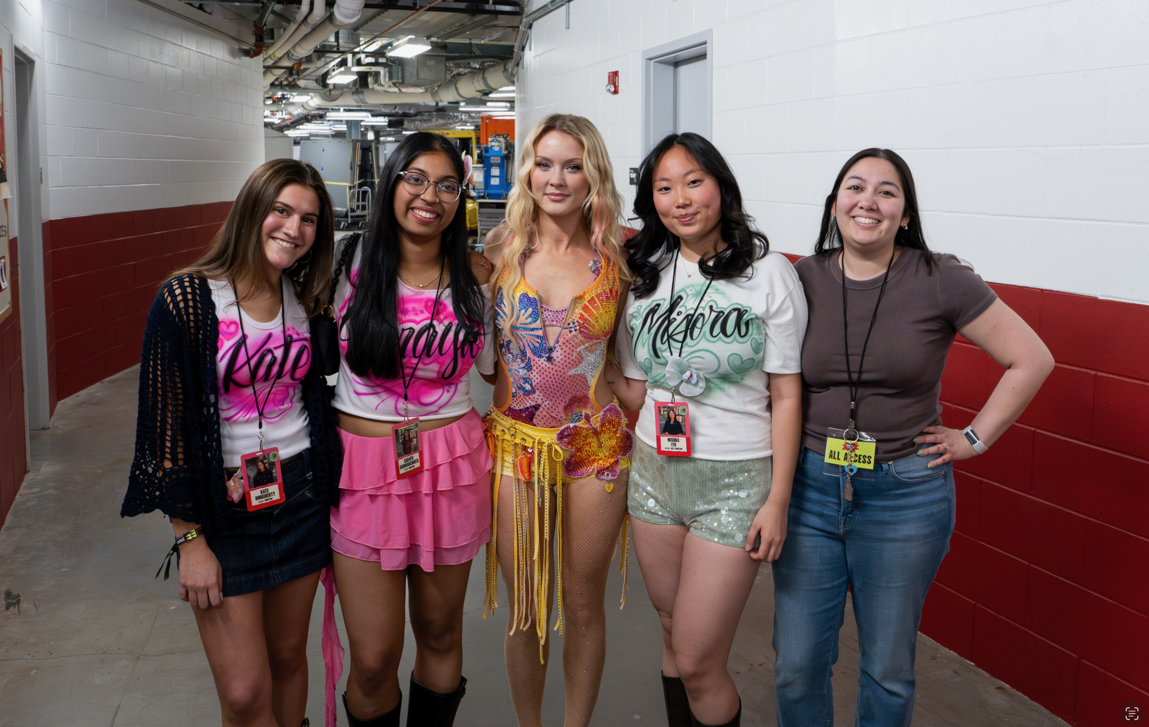 Five young women standing shoulder to shoulder in a hallway, smiling at the camera. The woman in the center is dressed in a colorful costume with feathers and sequins, while the others wear casual clothes with name tags and lanyards.