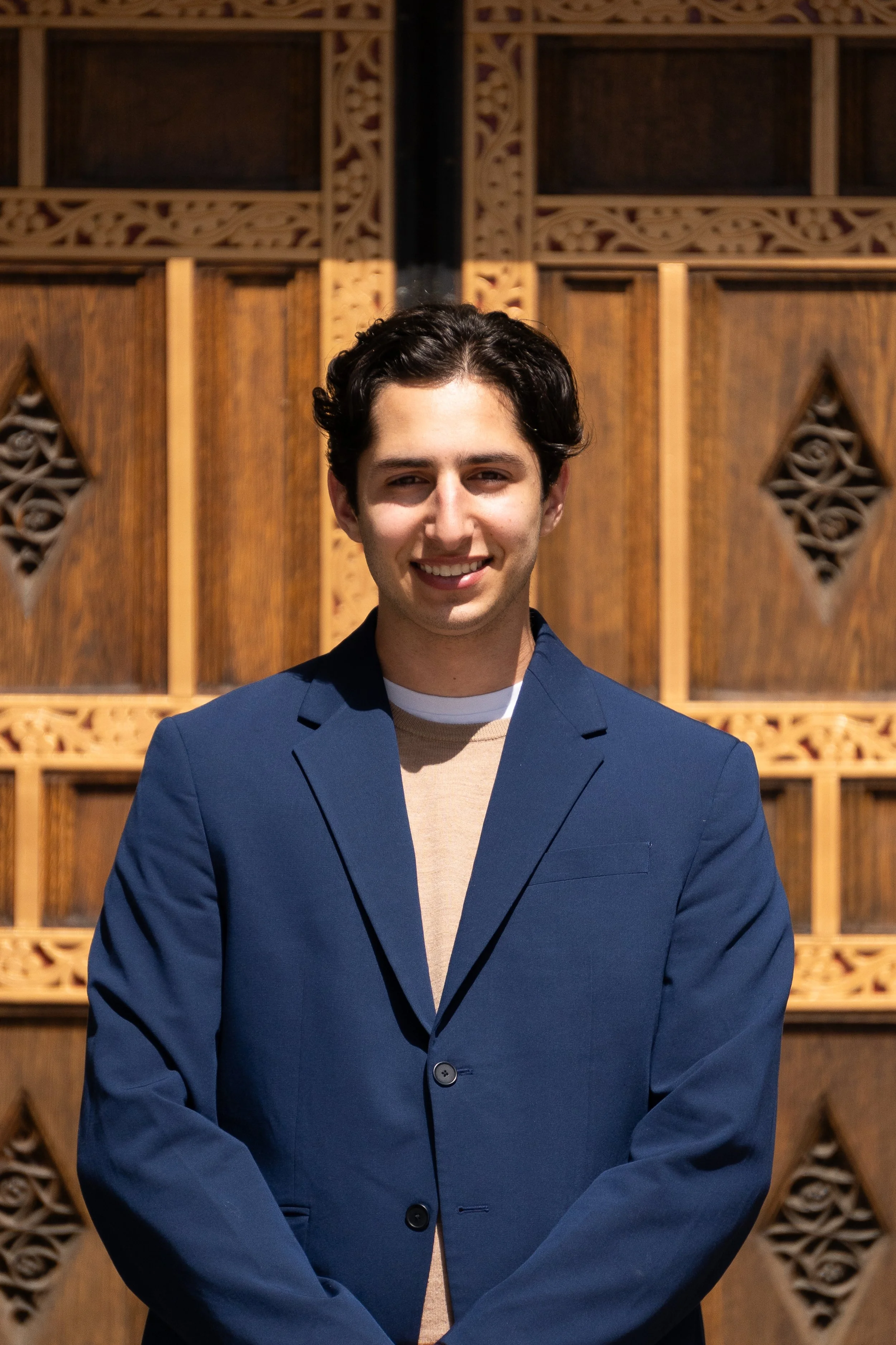Young man with dark wavy hair, smiling, wearing a blue blazer and light-colored shirt, standing in front of a decorative wooden background.