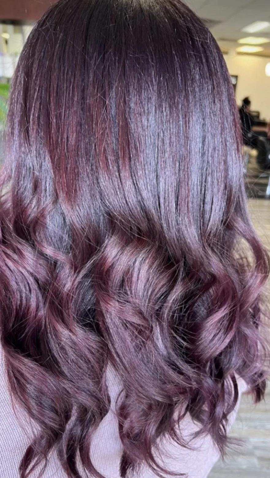 A person's hair styled in loose curls with a gradient color from dark at the roots to a pinkish-purple toward the ends.