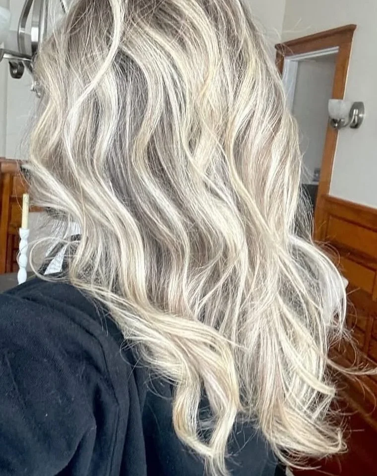 A person with long, wavy platinum blonde hair, sitting in a room with wooden furniture and a mirror.