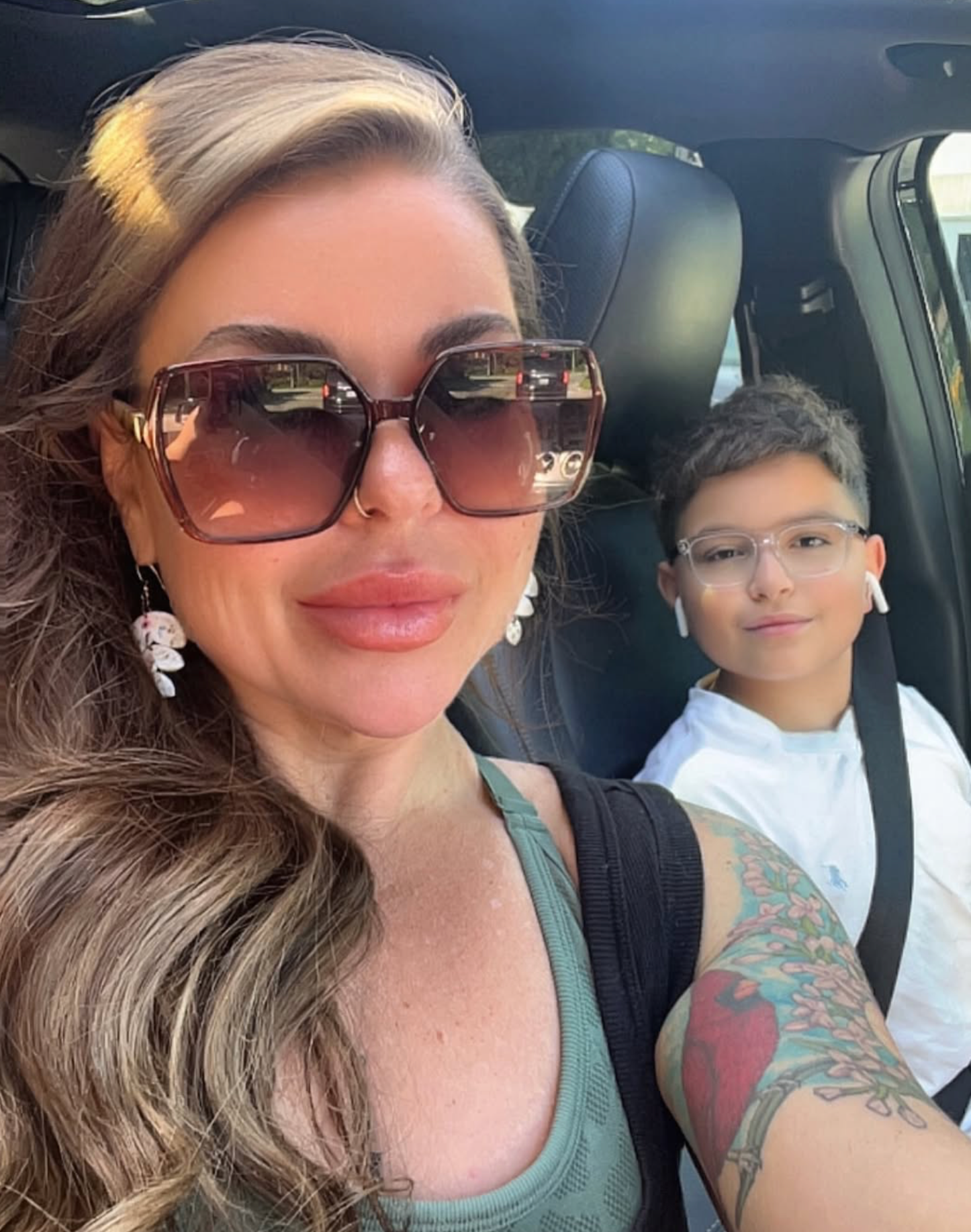 A woman with sunglasses and earrings taking a selfie with a young boy inside a car. The woman has long hair, a tattoo on her arm, and is wearing a tank top. The boy has short hair, glasses, and is wearing earbuds and a white T-shirt.