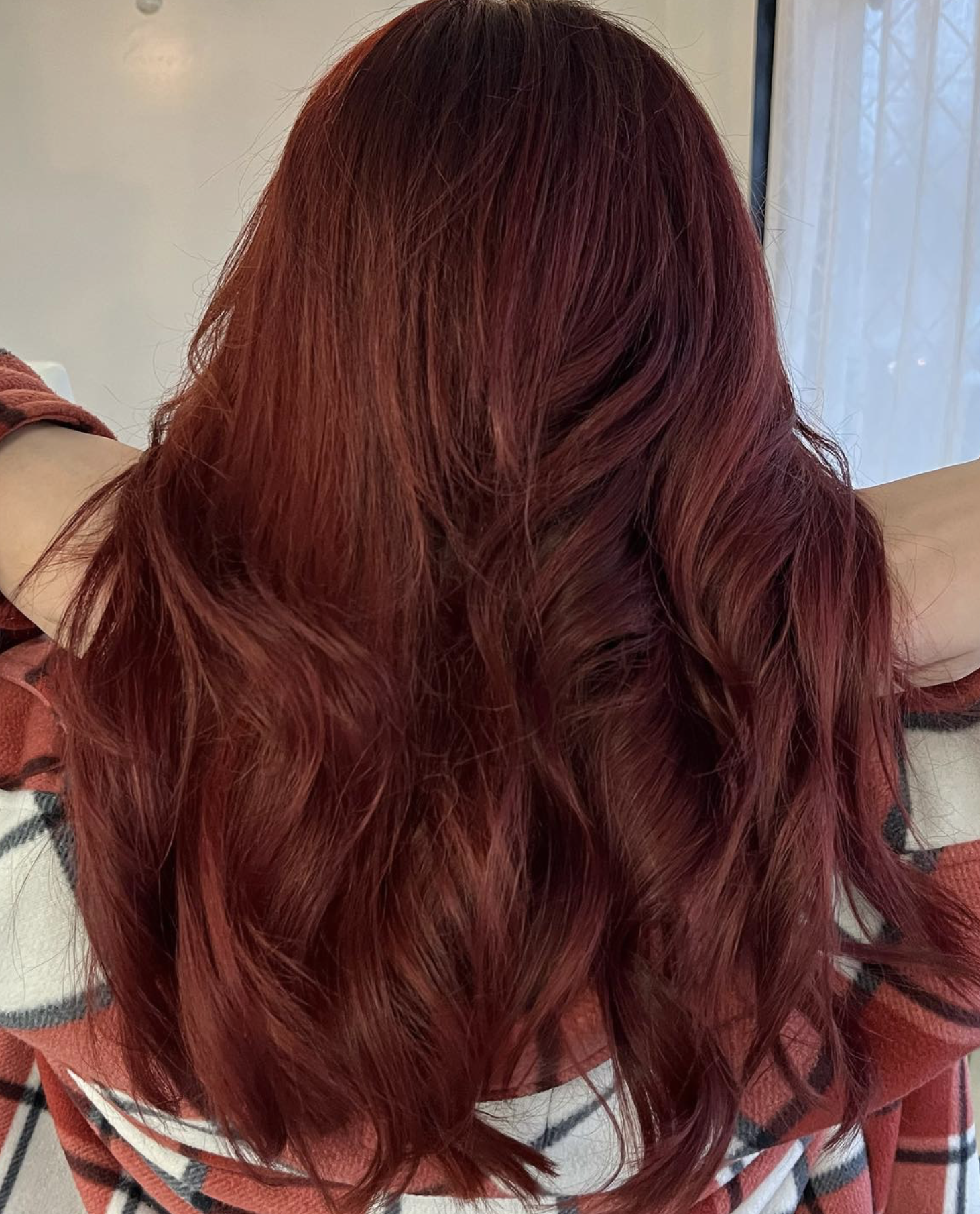 Person with long, wavy, red hair, sitting with arms behind head, indoors near a window with sheer curtains.