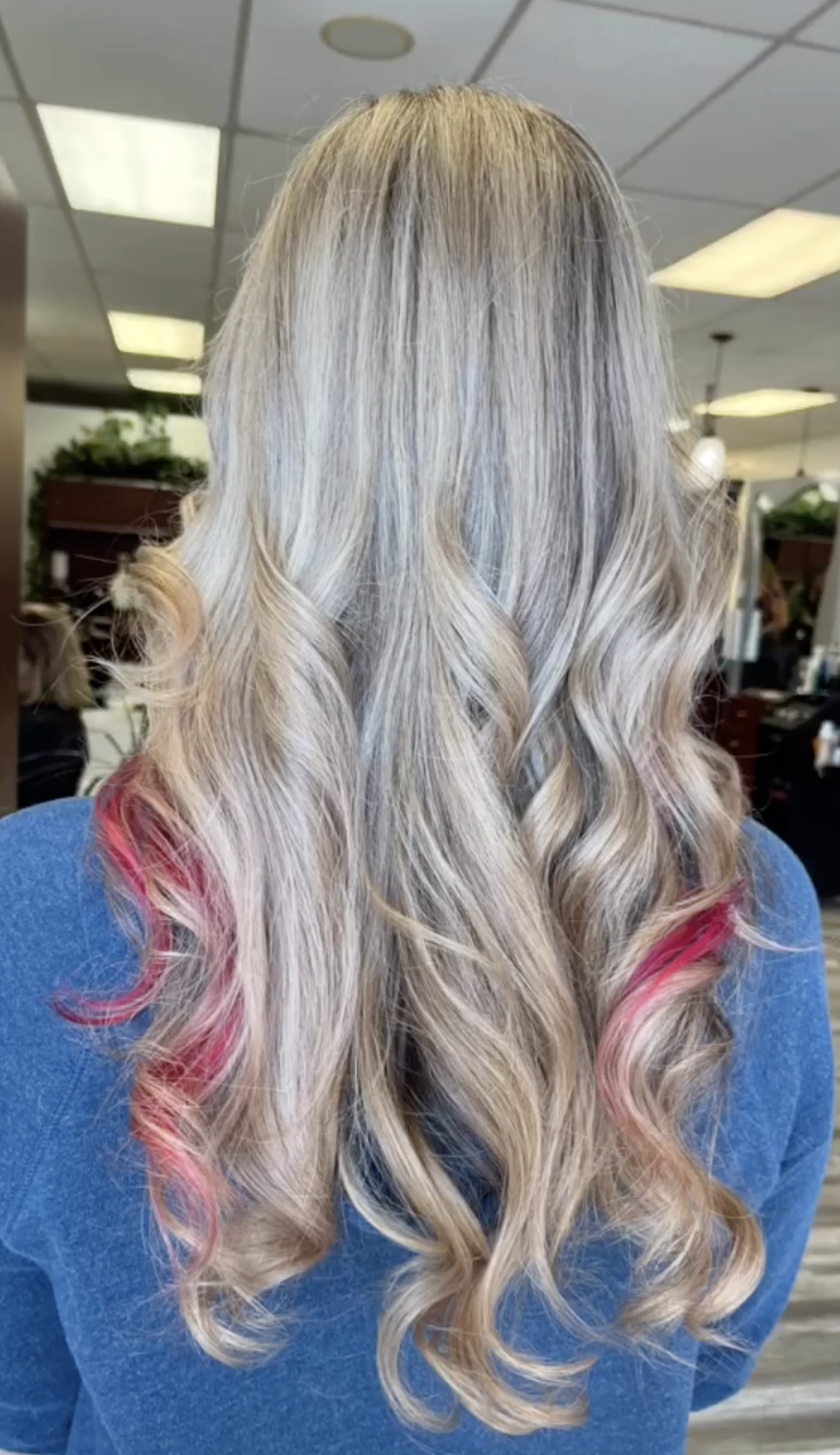 Back view of a woman with long, wavy, blonde hair with pink tips, sitting in a salon.