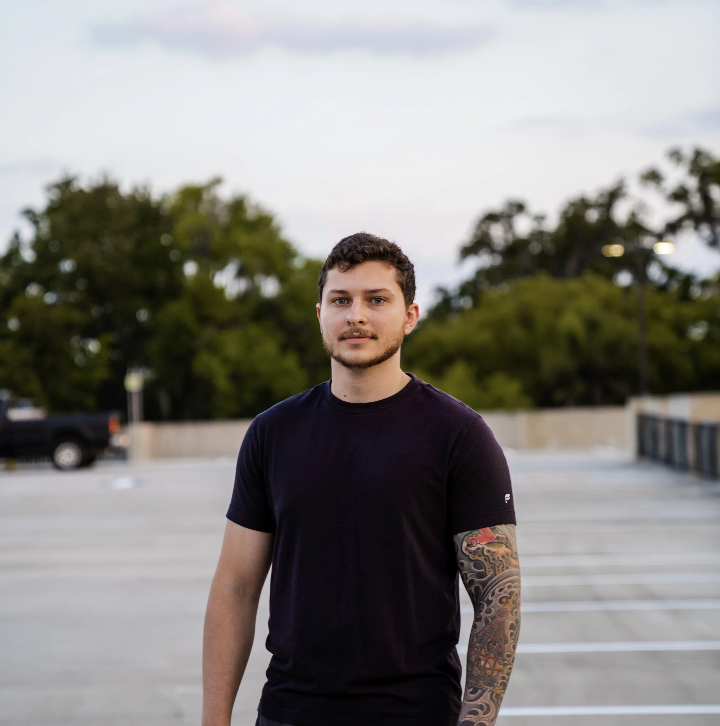 Young man with short dark hair and a tattoo sleeve on his left arm, wearing a black t-shirt, standing in an empty parking lot during dusk with trees and a cloudy sky in the background.