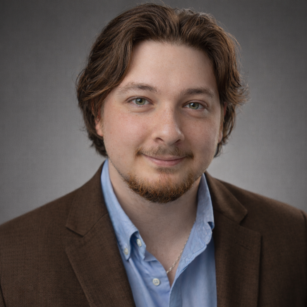 A young man with light skin, brown hair, and a goatee, wearing a brown blazer and a light blue collared shirt, smiling softly at the camera against a gray background.