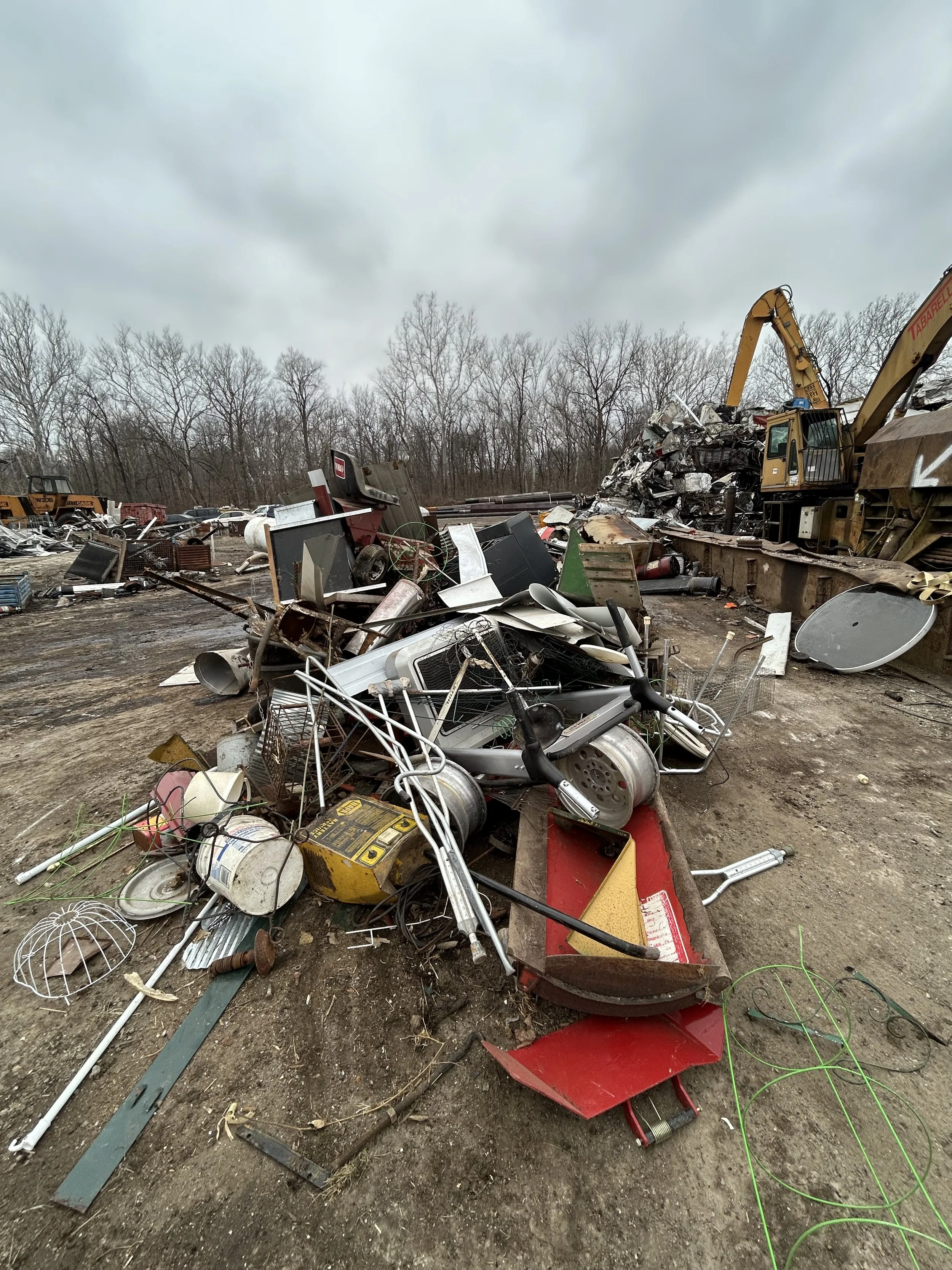 Pile of scrap metal and broken items in an outdoor scrapyard with overcast sky and leafless trees in the background.