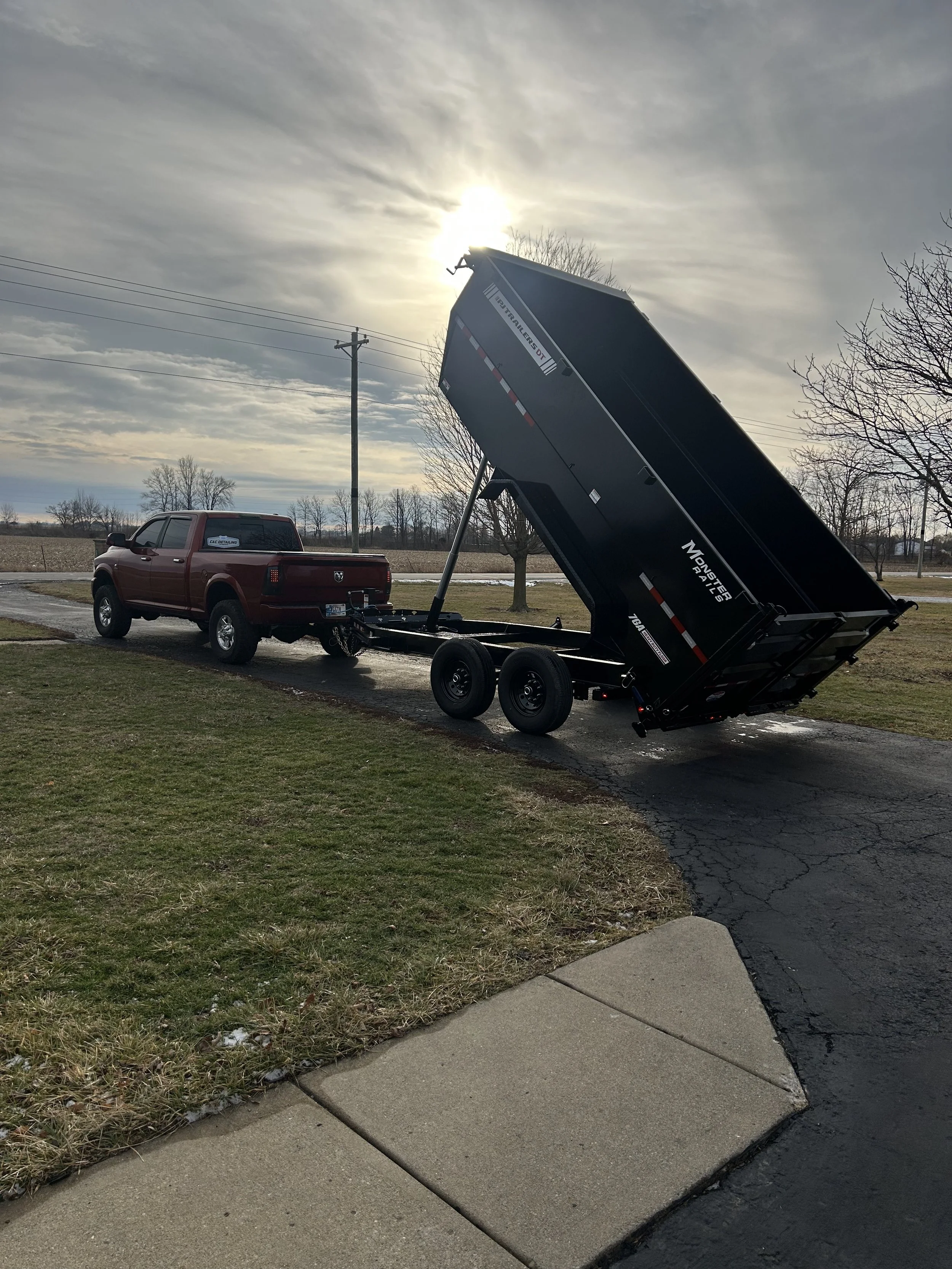 Red pickup truck with a black dump trailer tilted upward in a parking lot on a cloudy day.