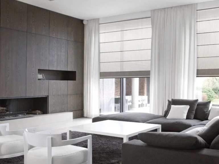 Modern living room with large windows, white curtains, gray sectional sofa with pillows, white coffee table, white and gray chairs, and a wooden accent wall.