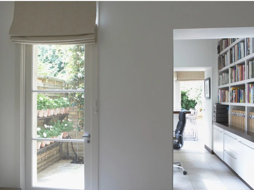 A white door with glass panes leading to a small outdoor patio with plants, next to a doorway opening into a bright home office with a large bookshelf and a black office chair.