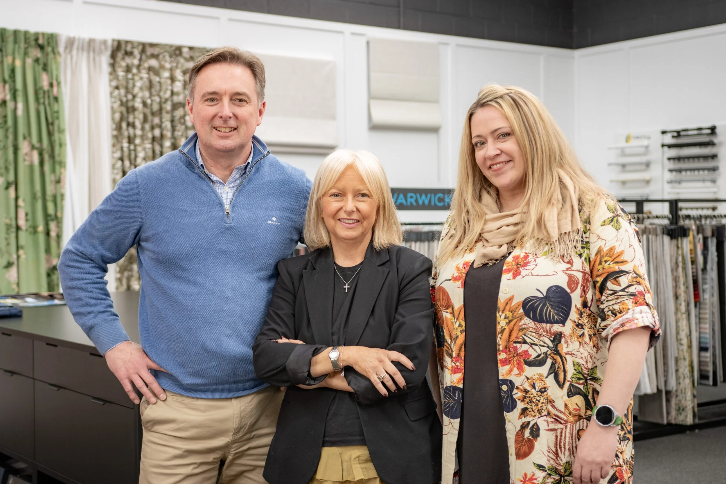 Three people standing together in a fabric and textile store, smiling at the camera. There are fabric bolts and sewing supplies in the background.