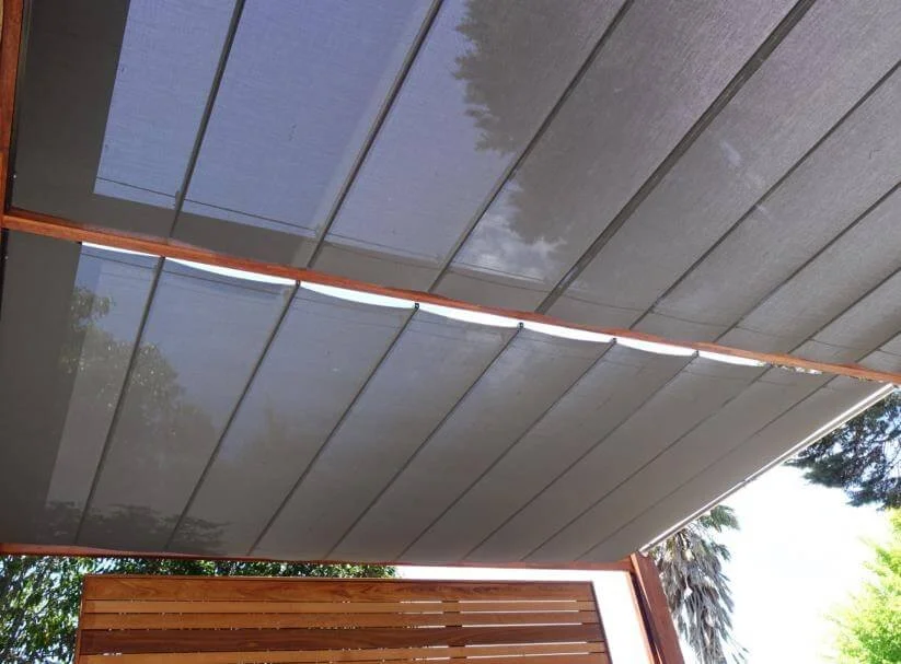 Close-up view of a modern outdoor patio ceiling with grey panels and wooden beams, with partial view of trees and sky in the background.