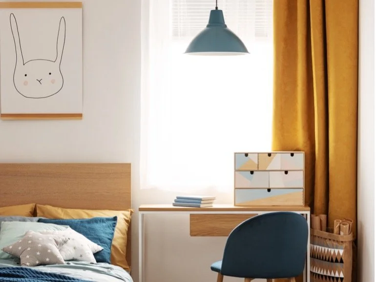 A cozy bedroom with a wooden bed frame, colorful pillows, a table drawer with a geometric design, a blue desk chair, and a wall art of a simplistic rabbit face, illuminated by natural light from a window with yellow curtains.