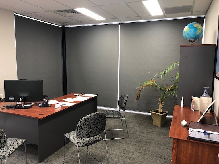 An empty office with a large L-shaped desk, two chairs, a potted plant, a filing cabinet with a globe on top, and a side table with a computer, papers, and a glass container.