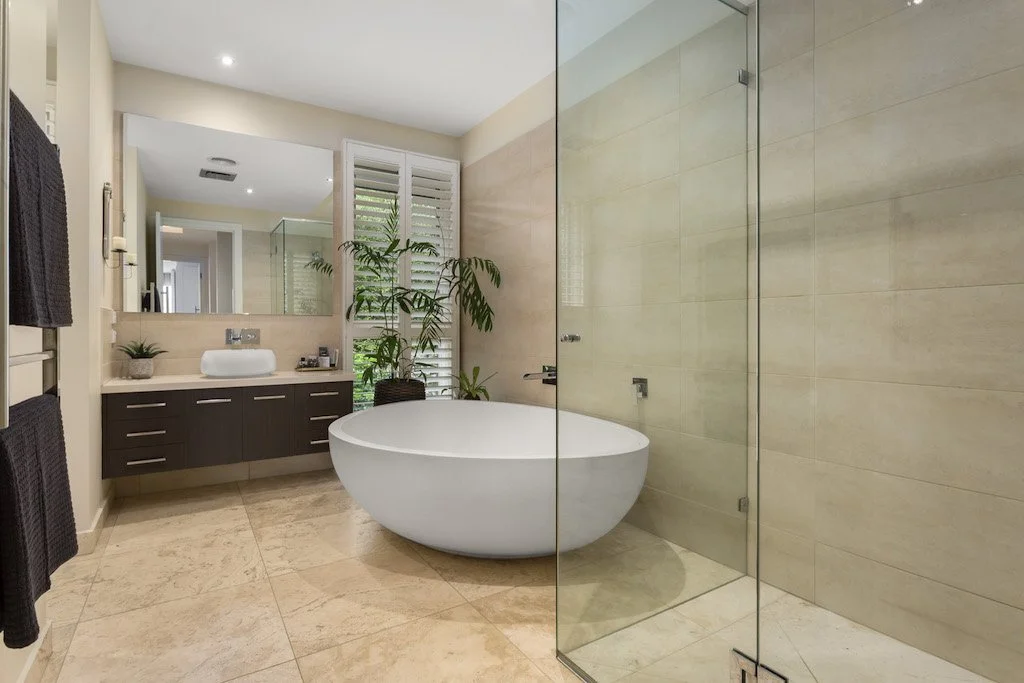 Modern bathroom with a white freestanding bathtub, a large mirror, a gray vanity with a sink, plants, and a glass-enclosed shower with beige tiles.