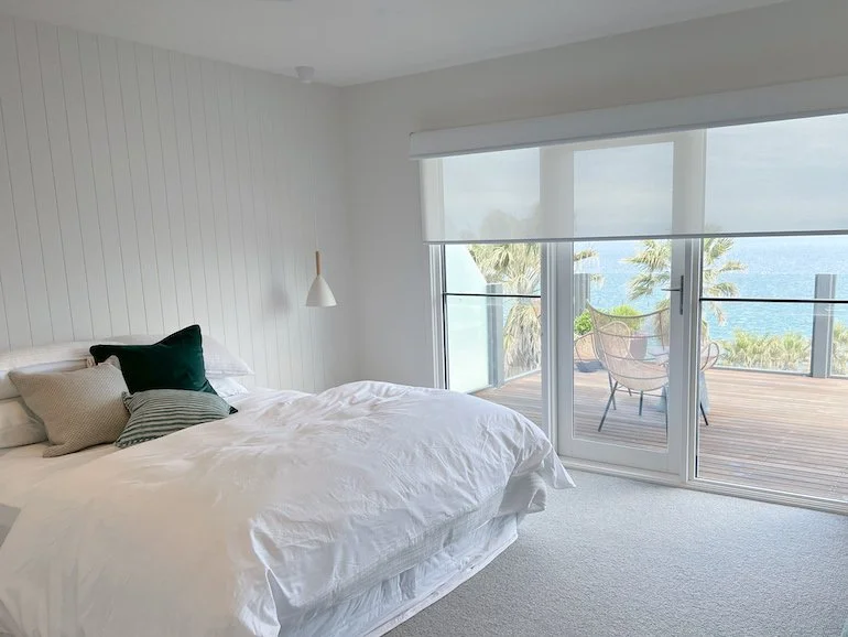 A bedroom with white bedding, a white wall, and a sliding glass door leading to a balcony with outdoor seating, overlooking palm trees and the ocean.