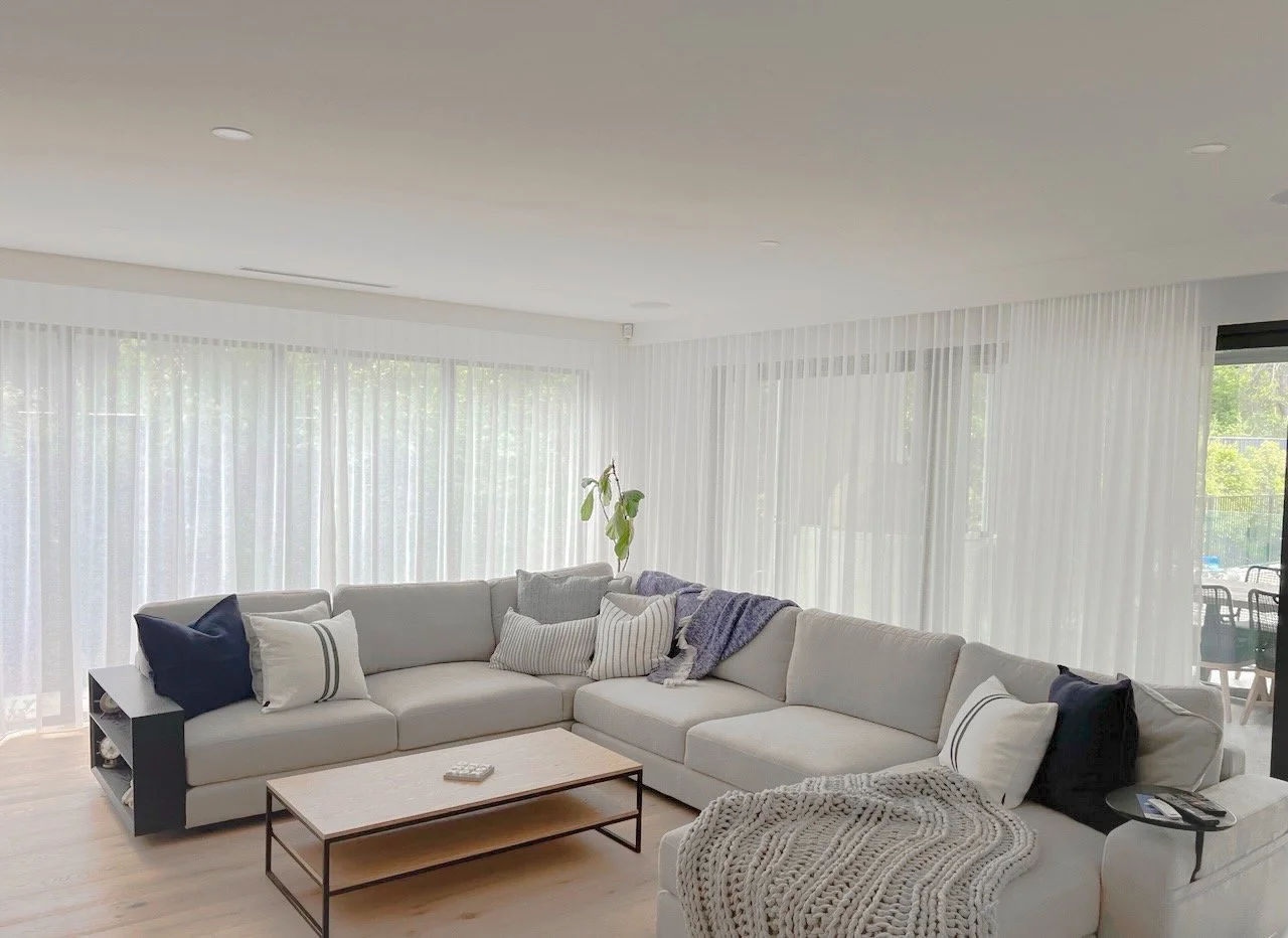 Bright living room with white sectional sofa, decorative pillows, a coffee table, sheer curtains, and a sliding glass door leading to an outdoor patio.