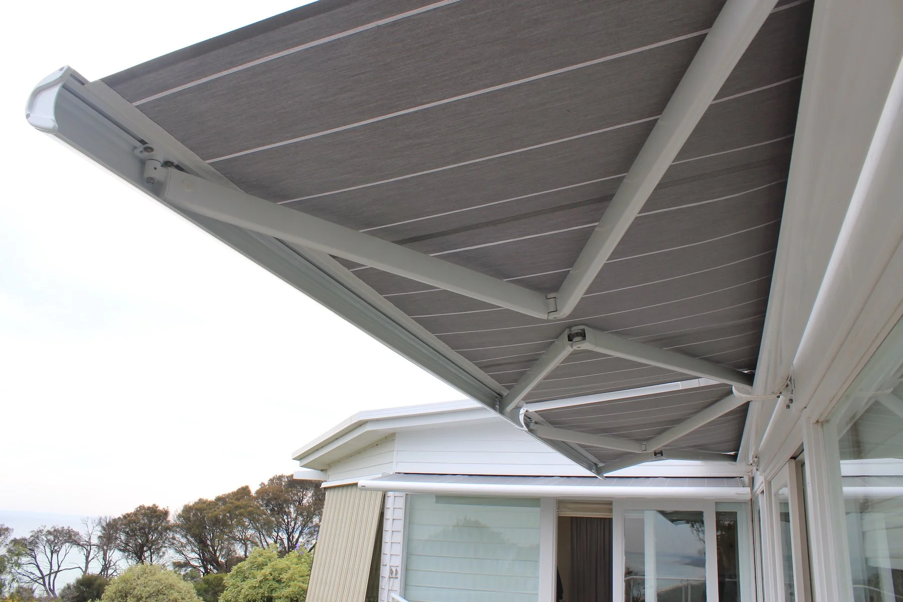Close-up view of a gray retractable awning extended above a house patio, with trees and sky in the background.